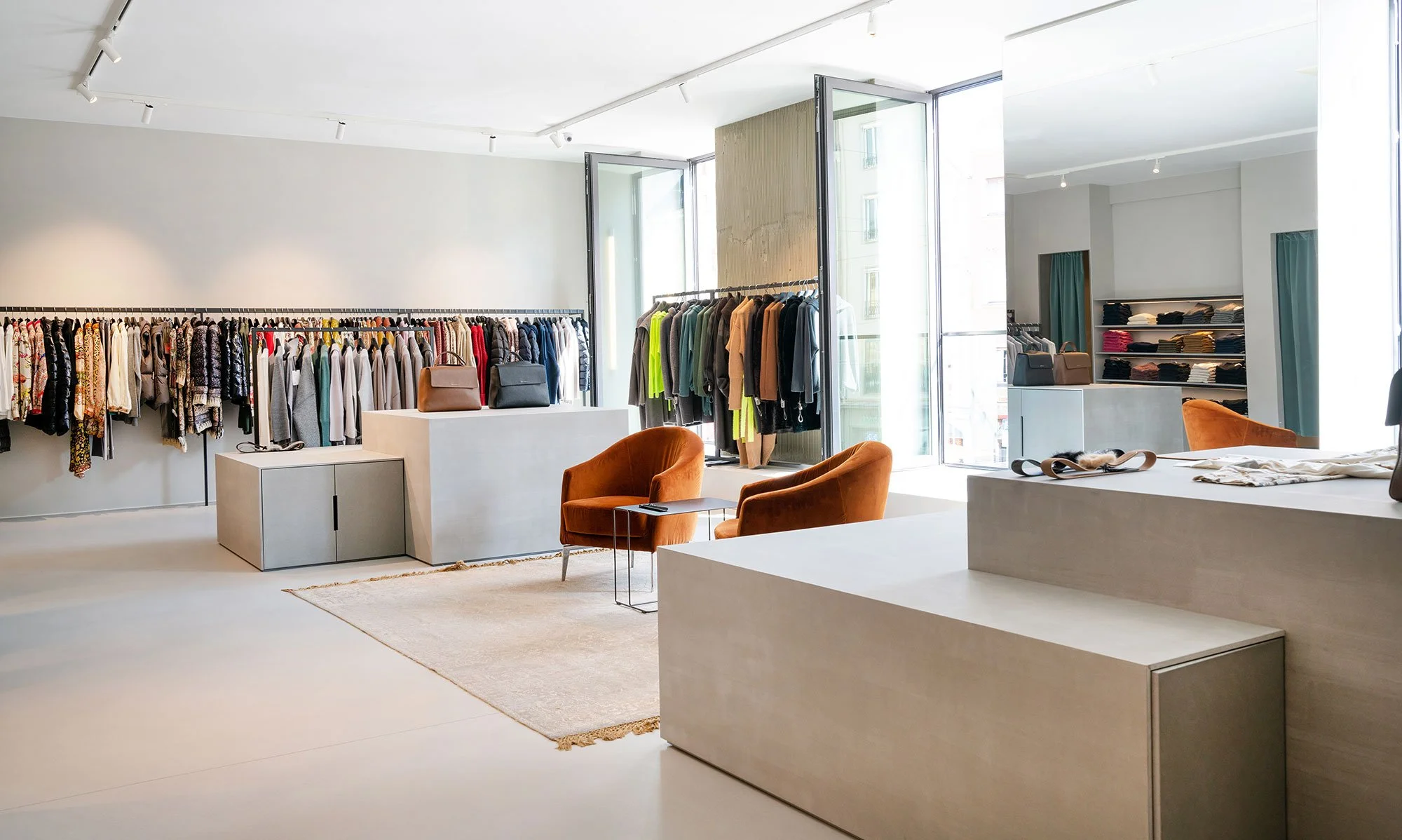 Ein Modegeschäft mit Kleidung auf Kleiderstangen, dekorativen Taschen auf Tischen, bequemen Sessel, Regal mit Stoffen und großer Fensterfront, Tageslicht im Innenraum.