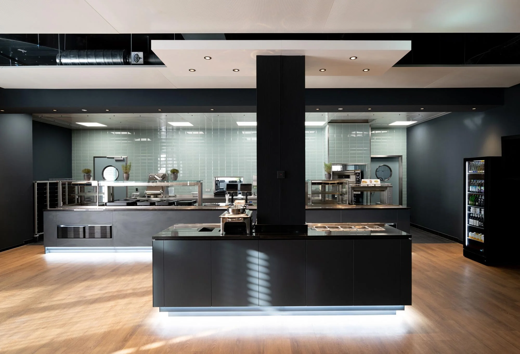 Moderne, leere Cafeteria mit schwarzem Tresen, hellgrüner Fliesenspiegel, Vorratskühlschrank, Pflanzen und Küchengeräten, Holzfußboden, dunkle Wände und Decke.