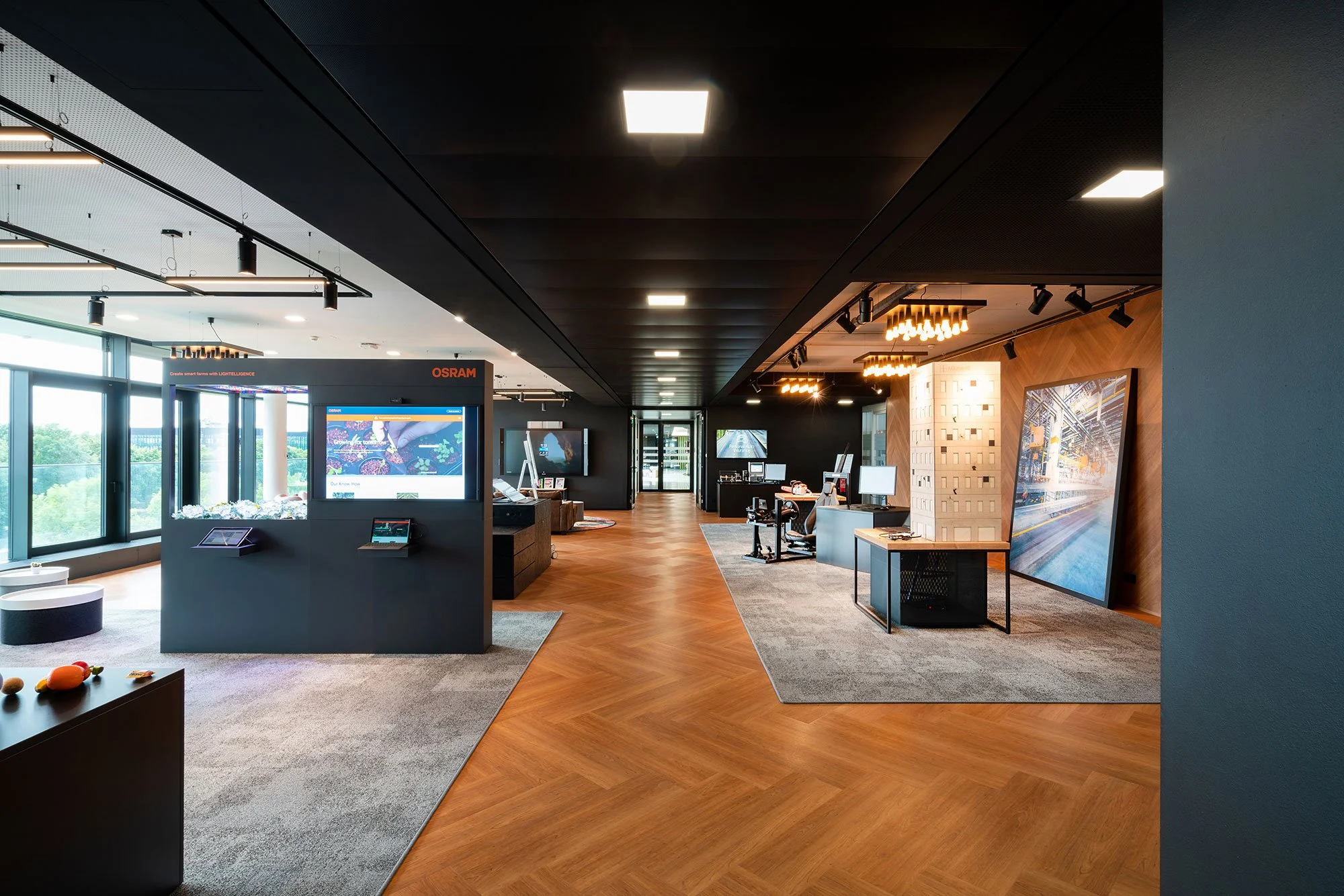 Innenansicht eines modernen Büros oder Showrooms mit dunklem Decke, großen Fenstern auf der linken Seite, einem Empfangstresen mit Bildschirmen und mehreren Arbeitsstationen in warmem Holzdesign, großer Wandbildschirm und moderner Beleuchtung.