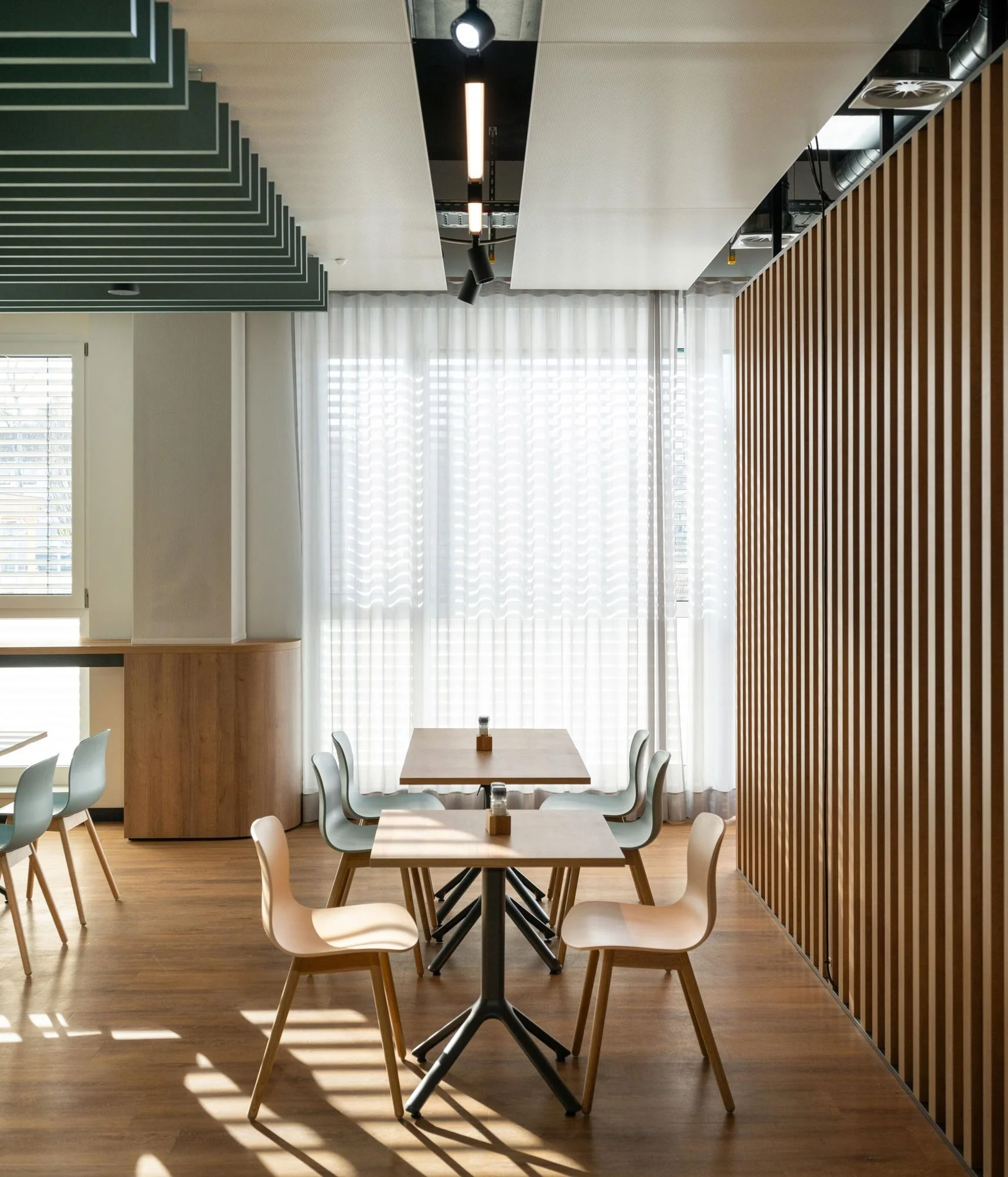 Innenraum eines modern eingerichteten Restaurants mit Holztischen, weißen Stühlen, großen Fenstern mit weißen Vorhängen, einer Holz- und einer schwarzen Trennwand sowie einer dezenten Beleuchtung an der Decke.