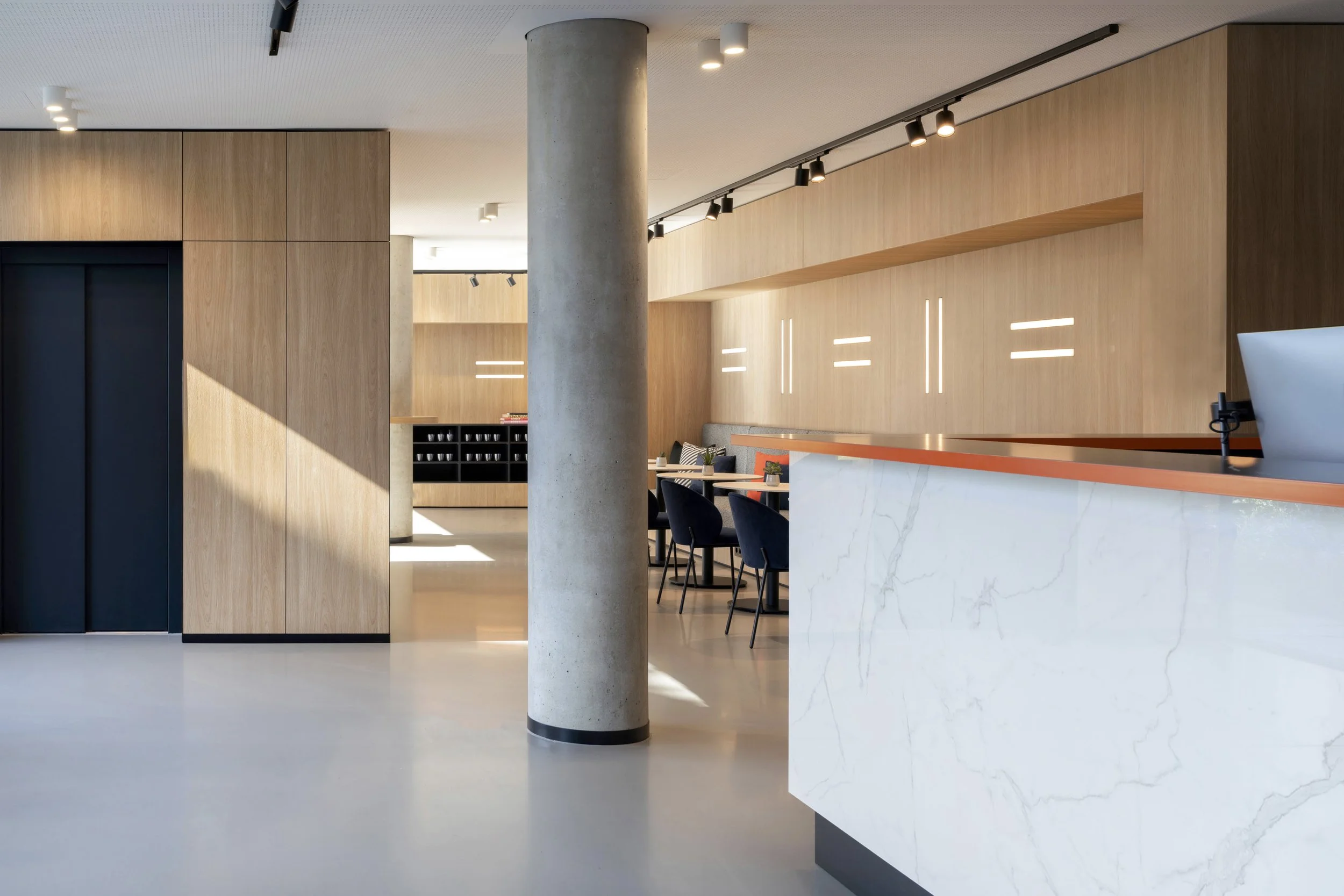 Moderner Empfangsbereich mit Holzverkleidungen, weißer Marmor-Rezeption und einer großen Betonstützsäule in einem Gebäude mit zeitgemäßem Design.