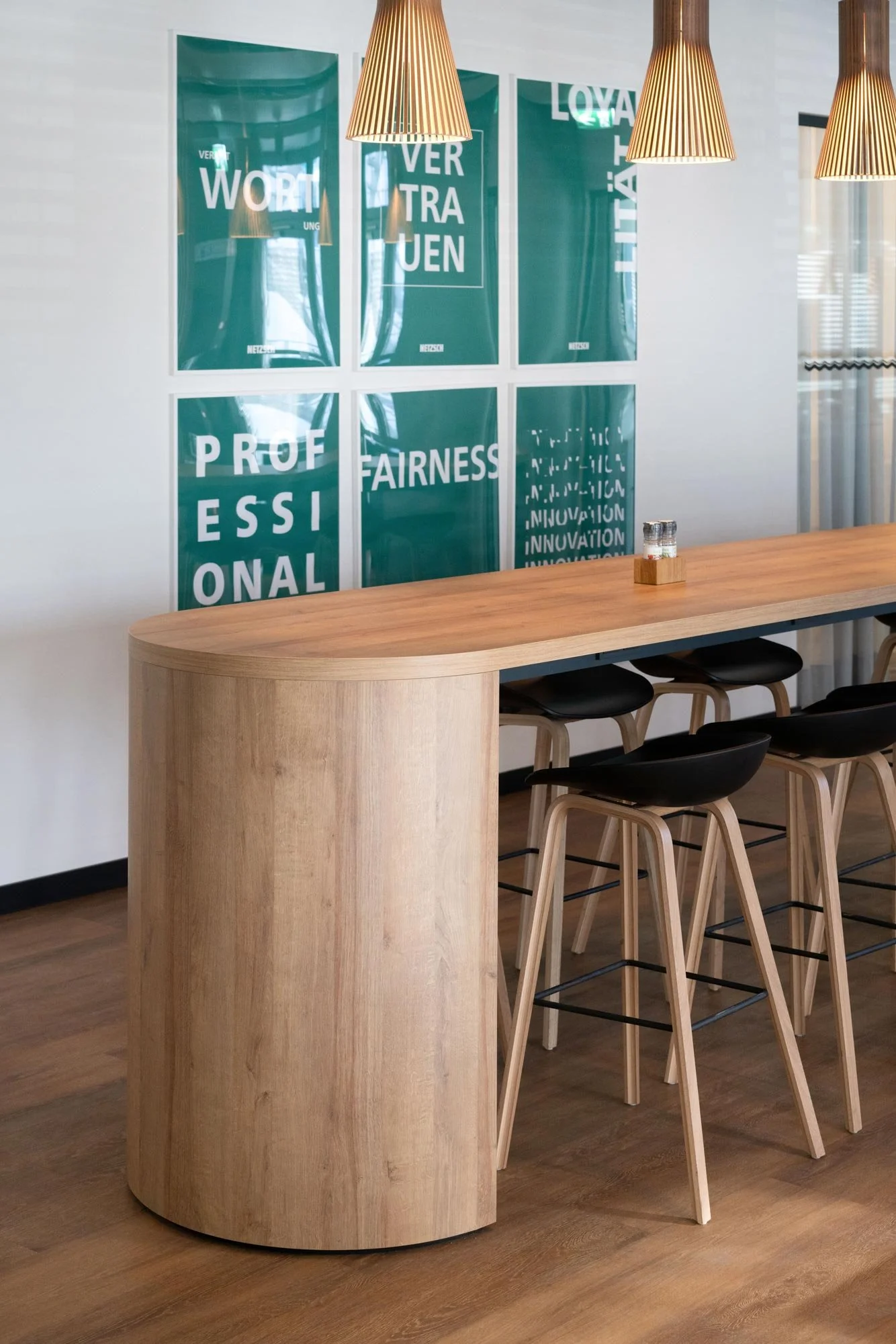 Ein modernes Café oder Bistro mit einem langen, runden Holzhochtisch, schwarzen Barhockern und einem Hintergrund mit grünen Wänden, auf denen Poster mit Texten wie "PROFESSIONAL" und anderen Worten in Weiß und Türkis hängen. Es gibt auch hölzerne Lam