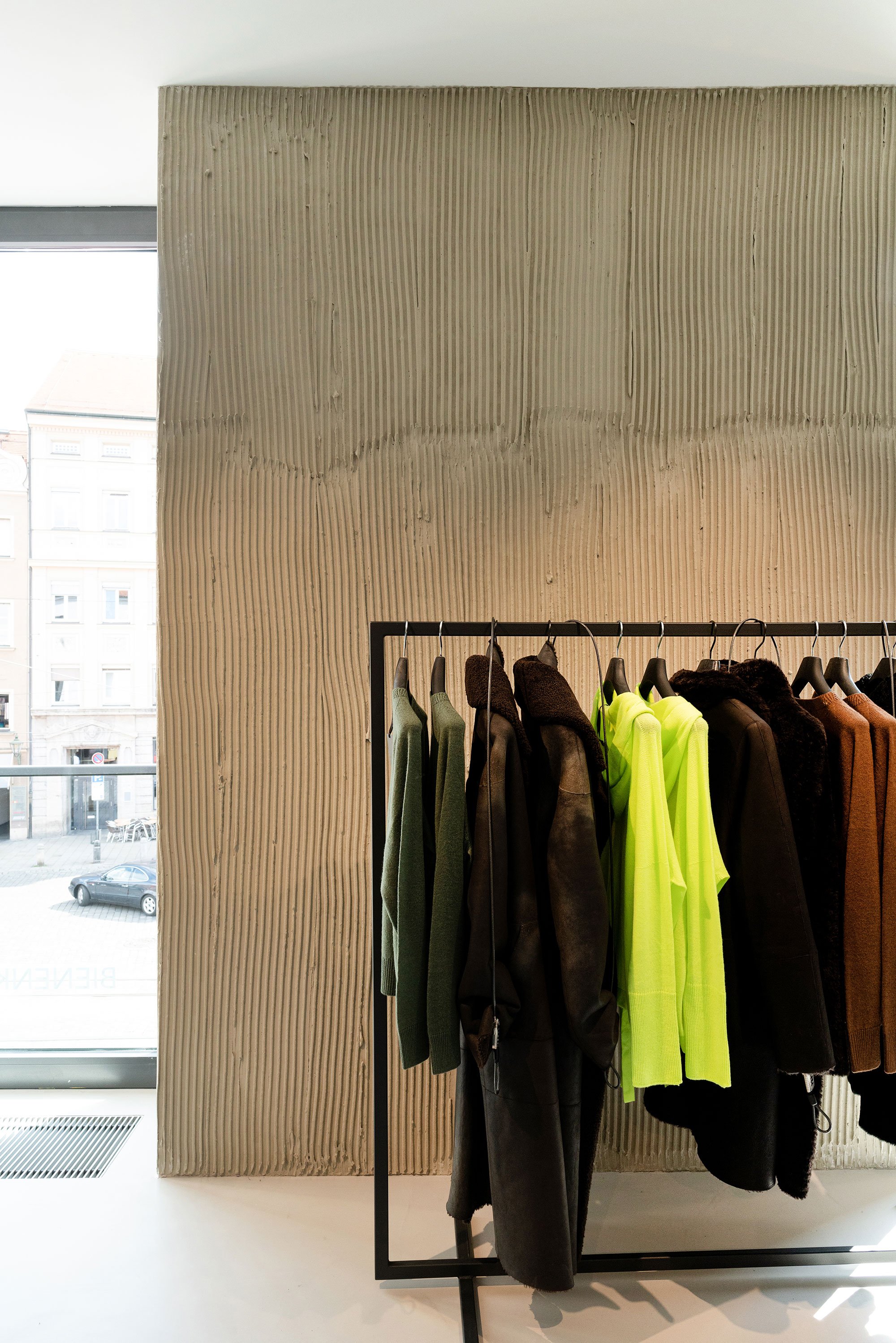 Kleiderständer mit mehreren Jacken und Mänteln in verschiedenen Farben, vor einem großen Fenster mit Blick auf die Stadt. Die Kleidung besteht aus dunklen, hellen und neonfarbenen Jacken, in einem Raum mit modernem, minimalistischen Design.