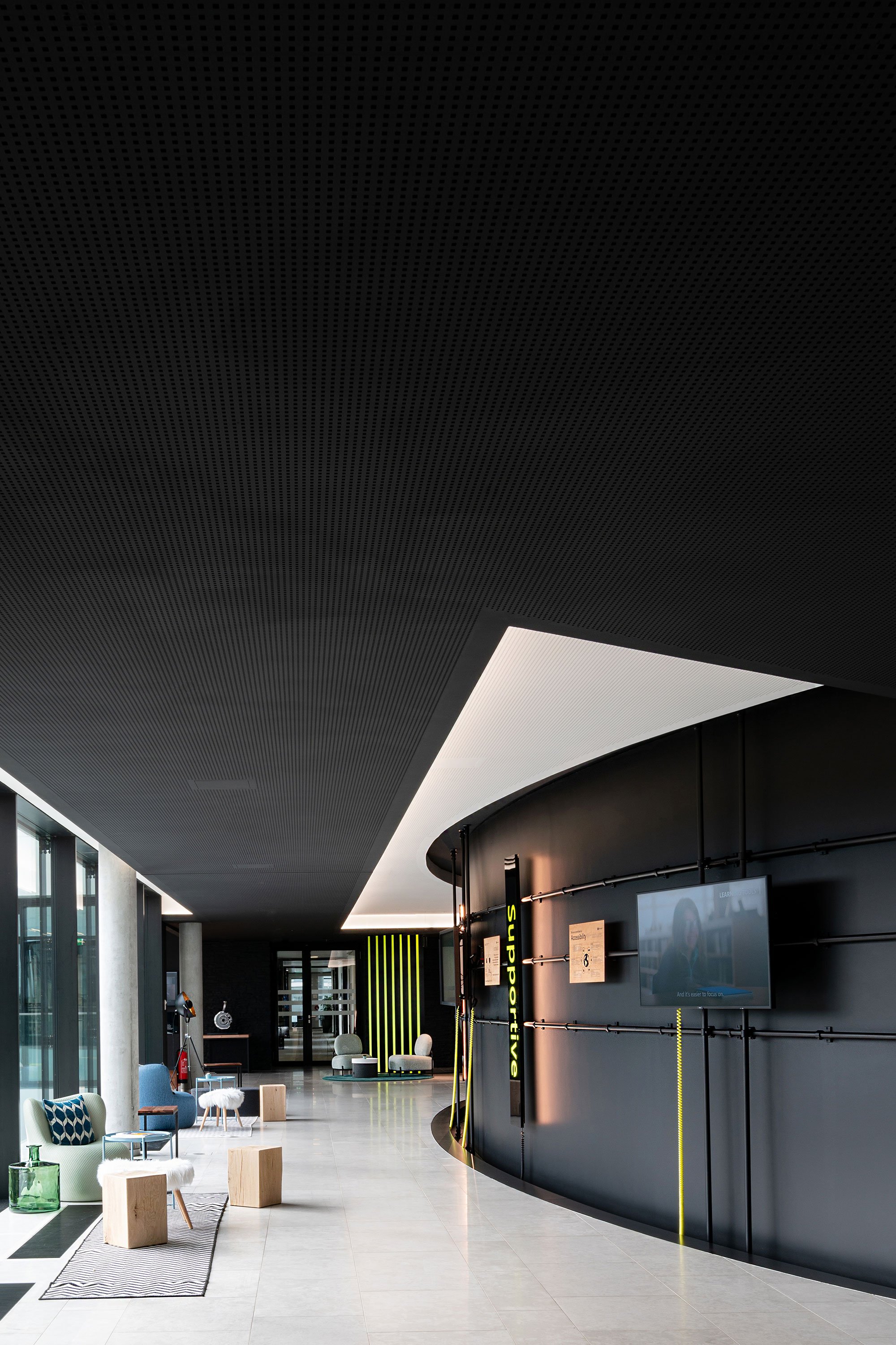Moderne Inneneinrichtung eines Wartezimmers mit bequemen Stühlen, Sofas und Couchtischen. Die Decke ist dunkel mit einer perforierten Struktur, und es gibt eine schwarze Wand mit einem Bildschirm und vertikalen Leuchtstreifen.