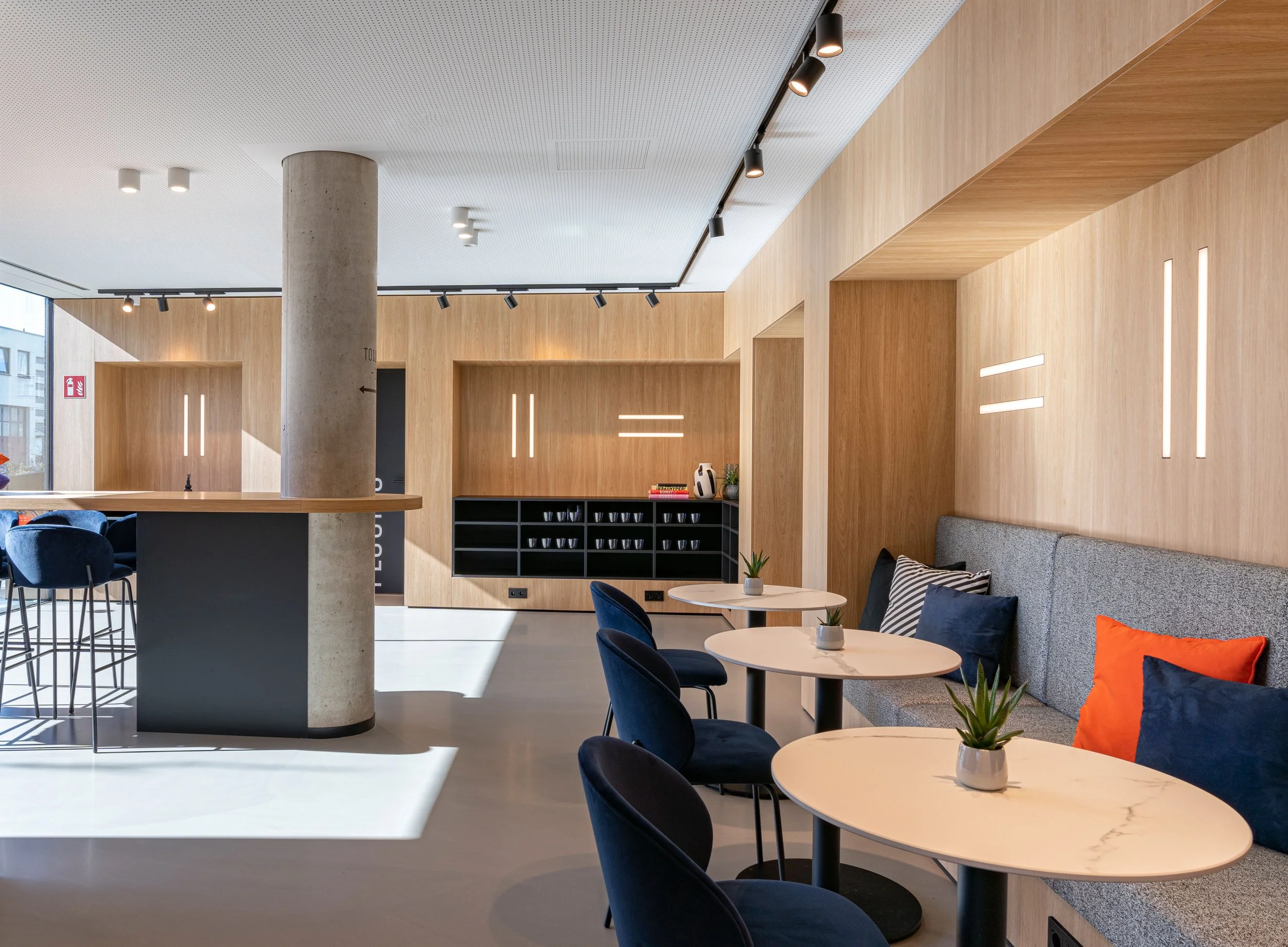 Innenraum eines modernen Café oder Lounge-Bereichs mit runden Tischen, blauen Sesseln, grauem Sofa, Holzverkleidungen und Deckenleuchten.