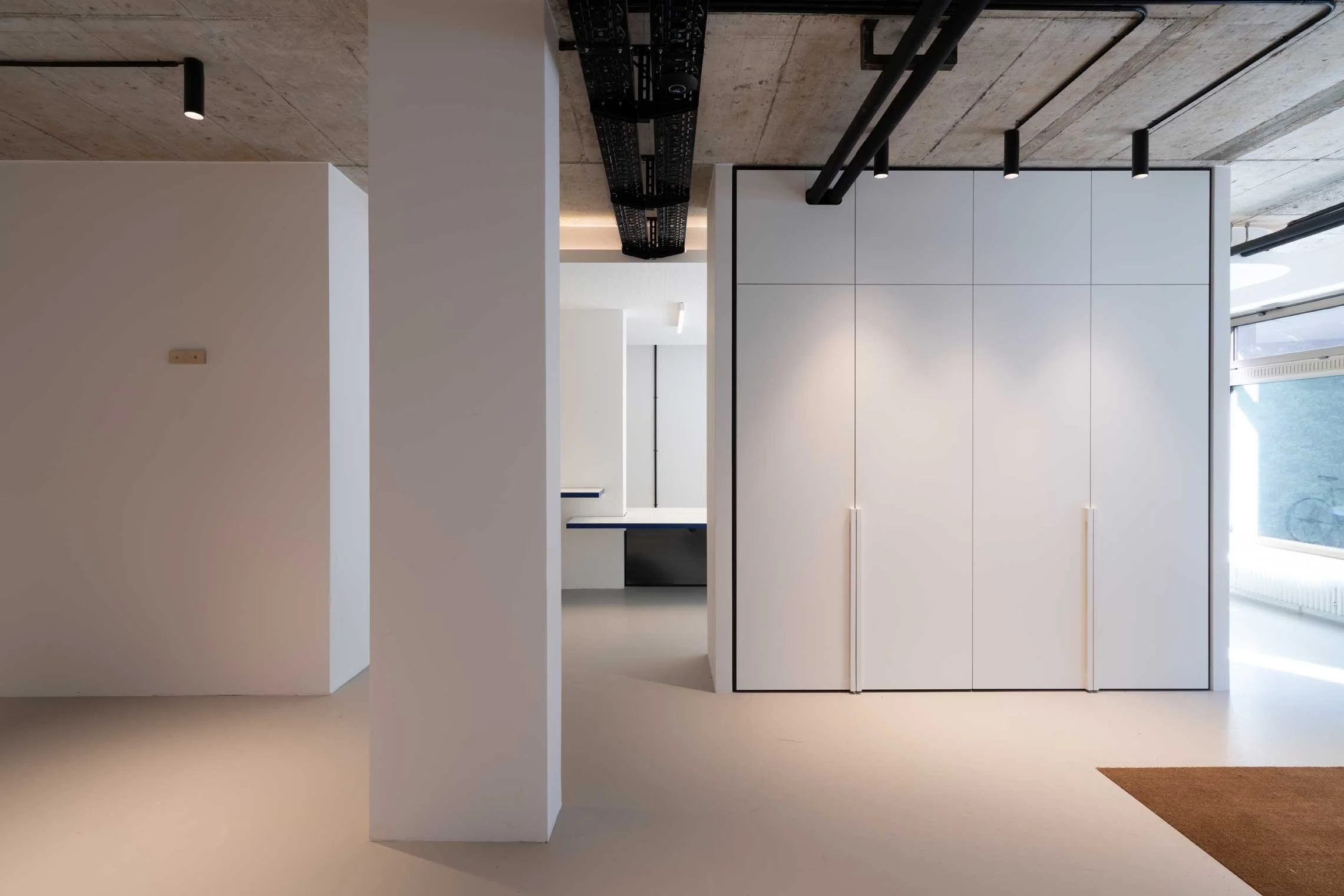 Innenraum eines modernen, minimalistischen Gebäudes mit weißen Wänden, großen Fenstern und sichtbaren schwarzen Rohrleitungen an der Decke.