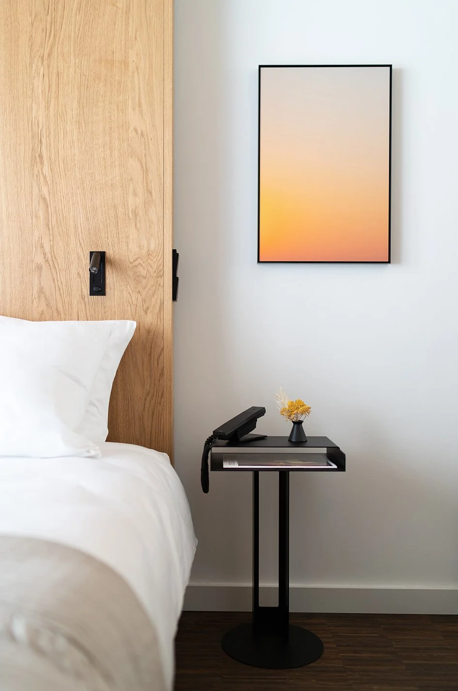 Ein Schlafzimmer mit einem weißen Bett, einem schwarzen Nachttisch, einem Wandbild mit einem orangefarbenen Sonnenuntergang, und einer kleinen Vase mit trockenen Blumen auf dem Nachttisch.