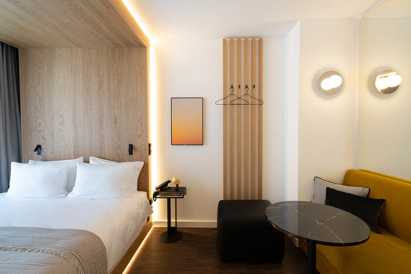 Modernes Hotelzimmer mit Doppelbett, Nachttisch, Wanddeko, Sitzbereich mit schwarzem Tisch und gelbem Sofa, warme Beleuchtung.