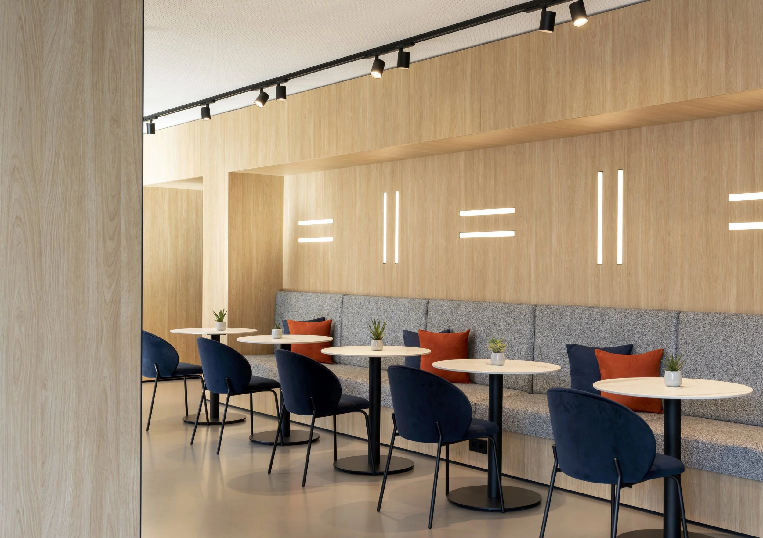 Ein moderner, heller Innenraum mit Holzvertäfelungen, Sitzgelegenheiten und kleinen Tischen, dekoriert mit kleinen Pflanzen, und moderner Beleuchtung.