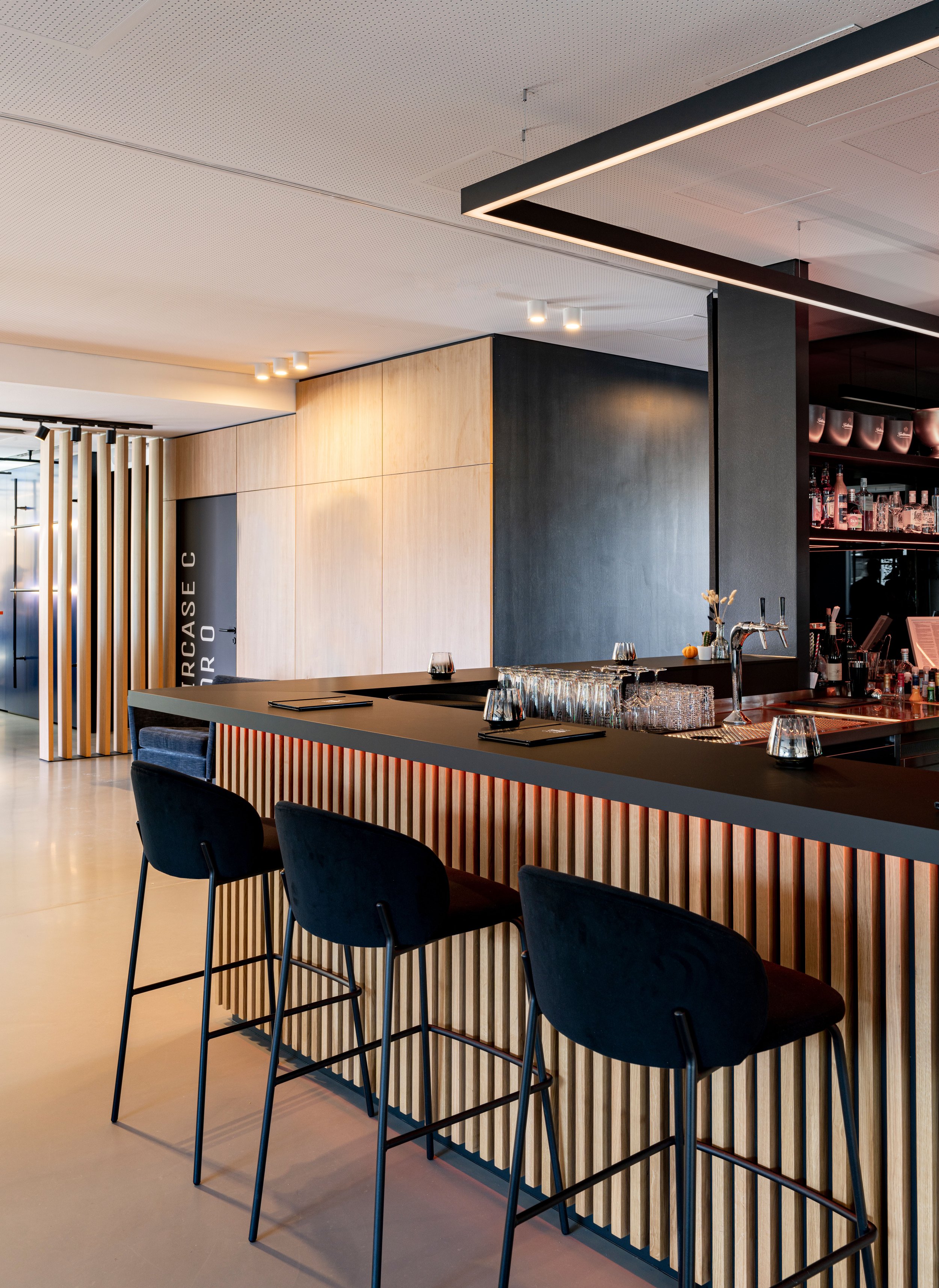 Innenansicht einer modernen Bar oder eines Restaurants mit schwarzem und hellem Holz-Dekor, hohem Thekenbereich mit schwarzen Barhockern, Wein- und Getränkespendern auf dem Bartresen, dezenten Beleuchtung und einer freundlichen Atmosphäre.