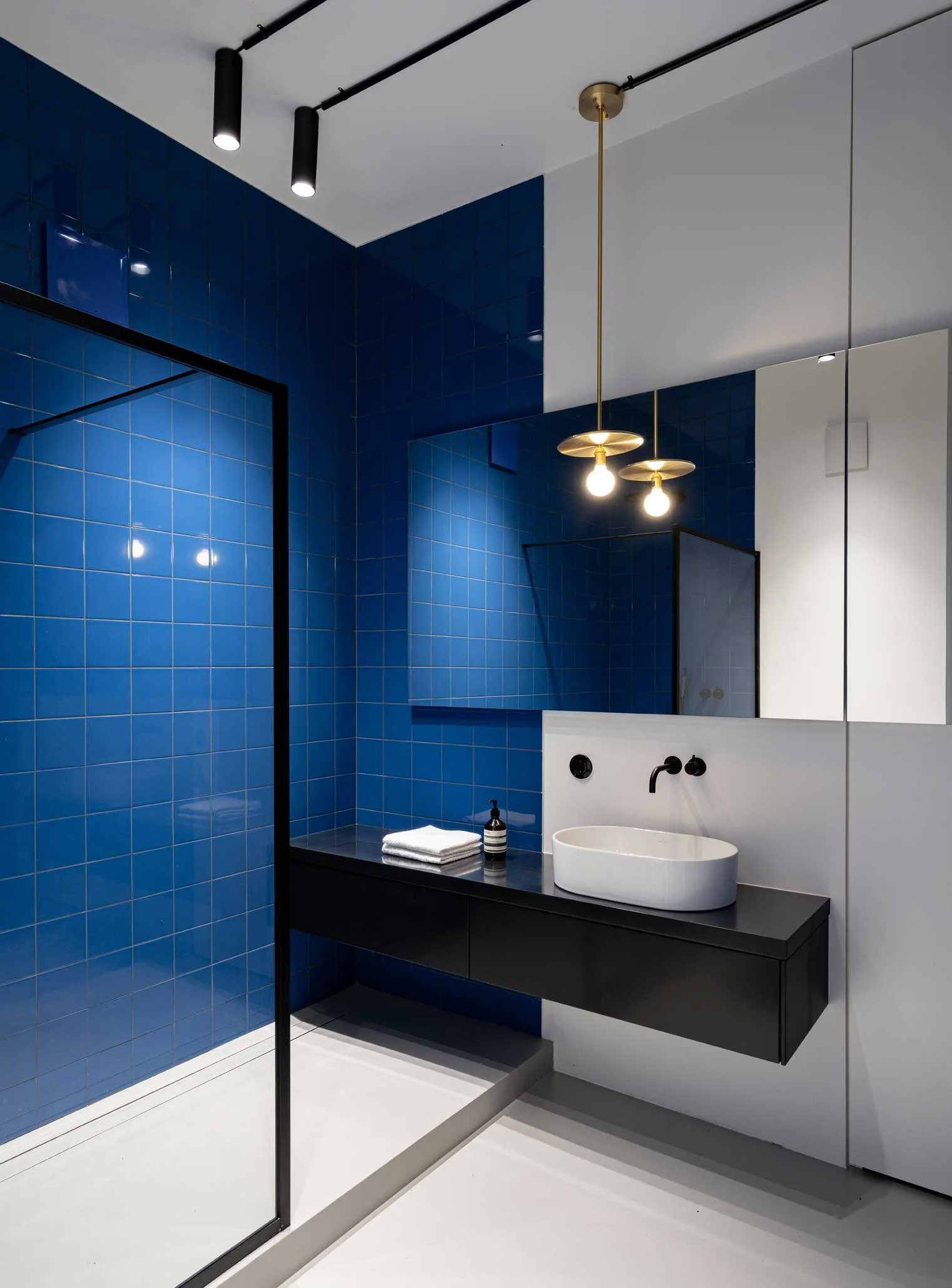 Moderner Badezimmer mit blauen Wandfliesen, weißen Wänden und einer schwarzen grauen Theke mit runden weißen Waschbecken. Es hängen dekorative Pendelleuchten, und im Spiegel ist ein Bereich des Badezimmers sichtbar.