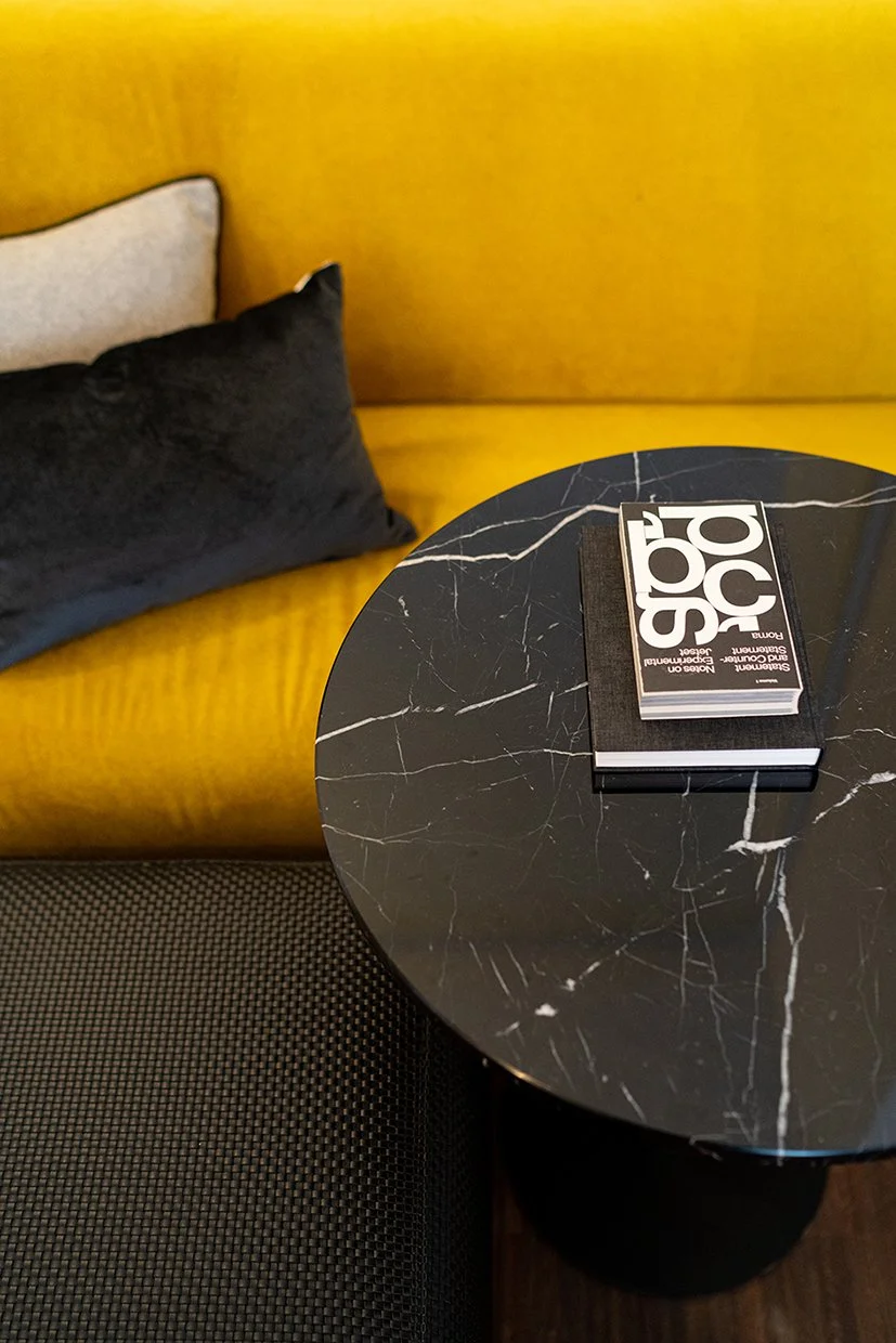 Ein schwarzer runder Tisch mit weißen Marmorlinien, auf dem ein schwarzes Notizbuch und ein Buch mit weißem und schwarzem Cover liegen, neben einer gelben Couch mit einem schwarzen und einem weißen Kissen.