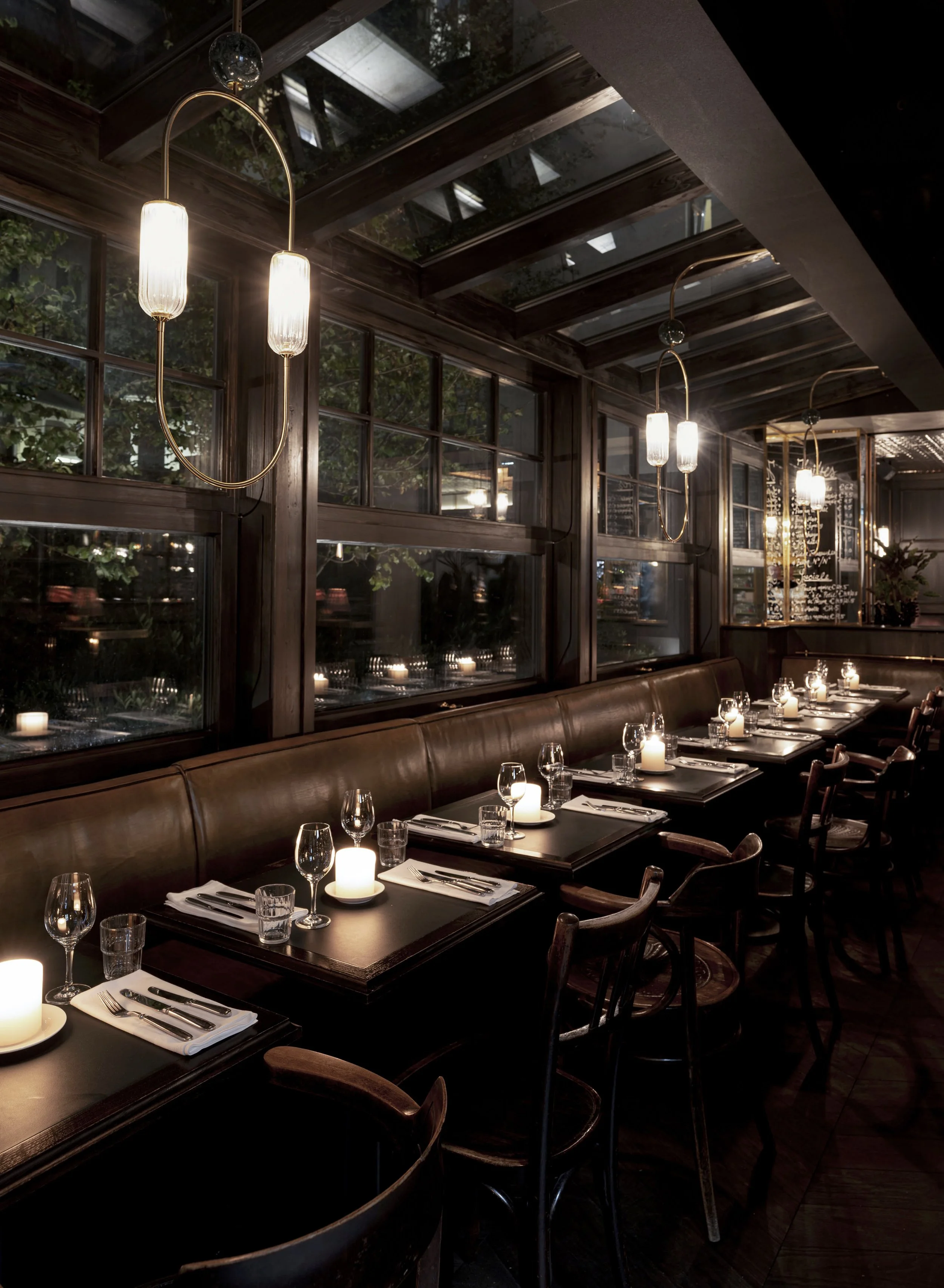 Ein eleganter, dunkler Restaurantraum mit Holzmöbeln, beleuchtet durch Kerzen auf den Tischen und schimmernden modernen Hängeleuchten. Große Fenster zeigen grünes Laub im Außenbereich.