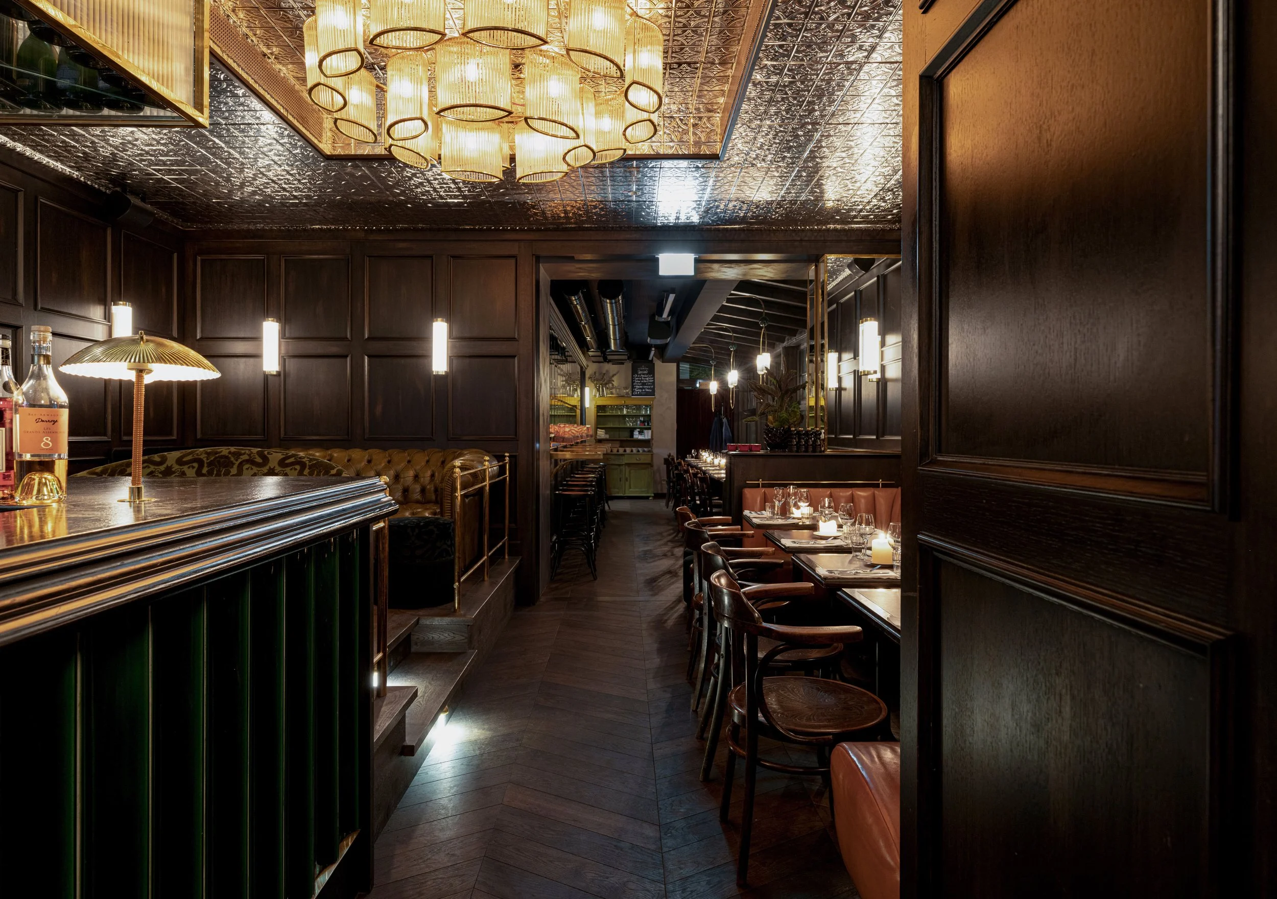 Ein stilvoll eingerichteter Restaurant- oder Barraum mit dunklen Holzvertäfelungen, einem dekorativen abgehängten Deckenpaneel mit Lichtinstallationen, Tischen mit Kerzen und bequemen Stühlen, eine gemütliche Atmosphäre.
