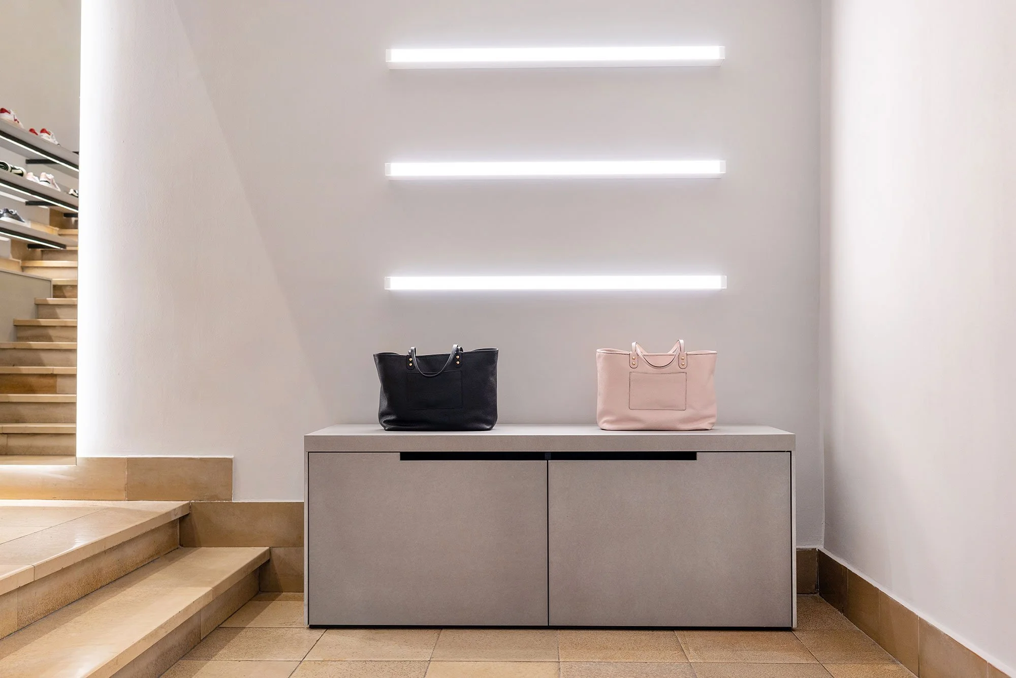 Zwei Handtaschen, eine schwarze und eine rosa, stehen auf einer modernen weißen Kommode vor einer weißen Wand mit horizontalen Beleuchtungslinien in einem eleganten Geschäft oder Eingangsbereich.