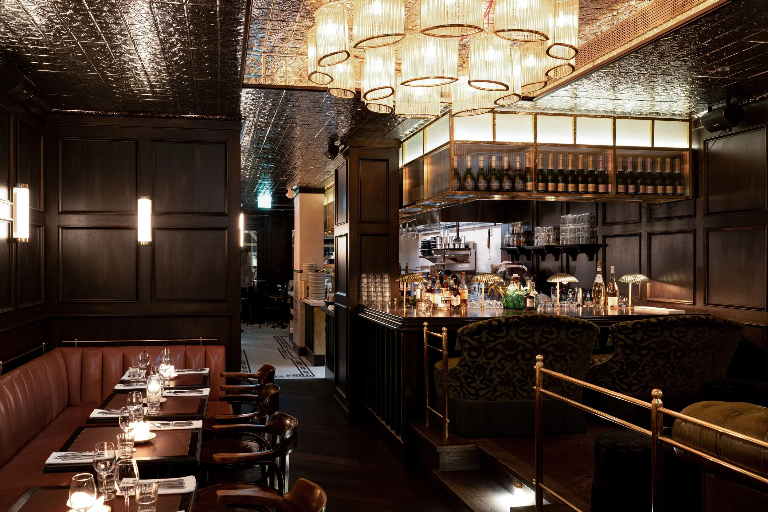 Ein elegantes Restaurant mit dunklem Holzinterieur, bequemen Sesseln, gedeckten Tischen mit Kerzen und einem stilvollen Barbereich, beleuchtet von großen, modernen Lampen an der Decke.
