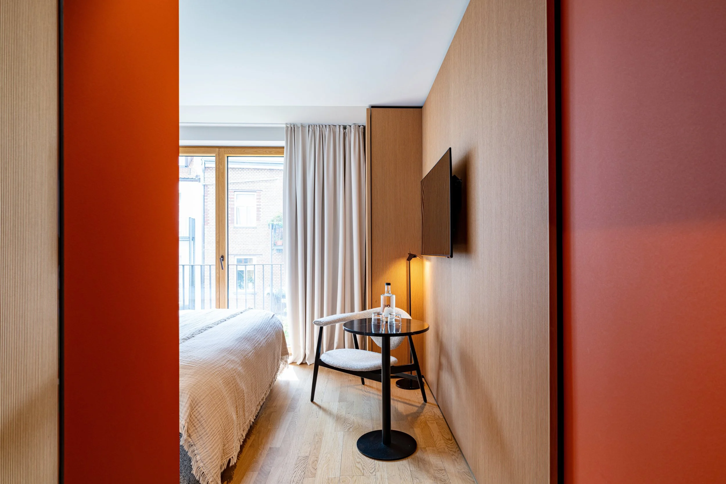 Blick in ein modernes Hotelzimmer mit Balkon, Bett, einem kleinen runden Tisch, zwei Stühlen, einem Flachbildfernseher an der Wand und weißen Vorhängen vor einem Fenster.