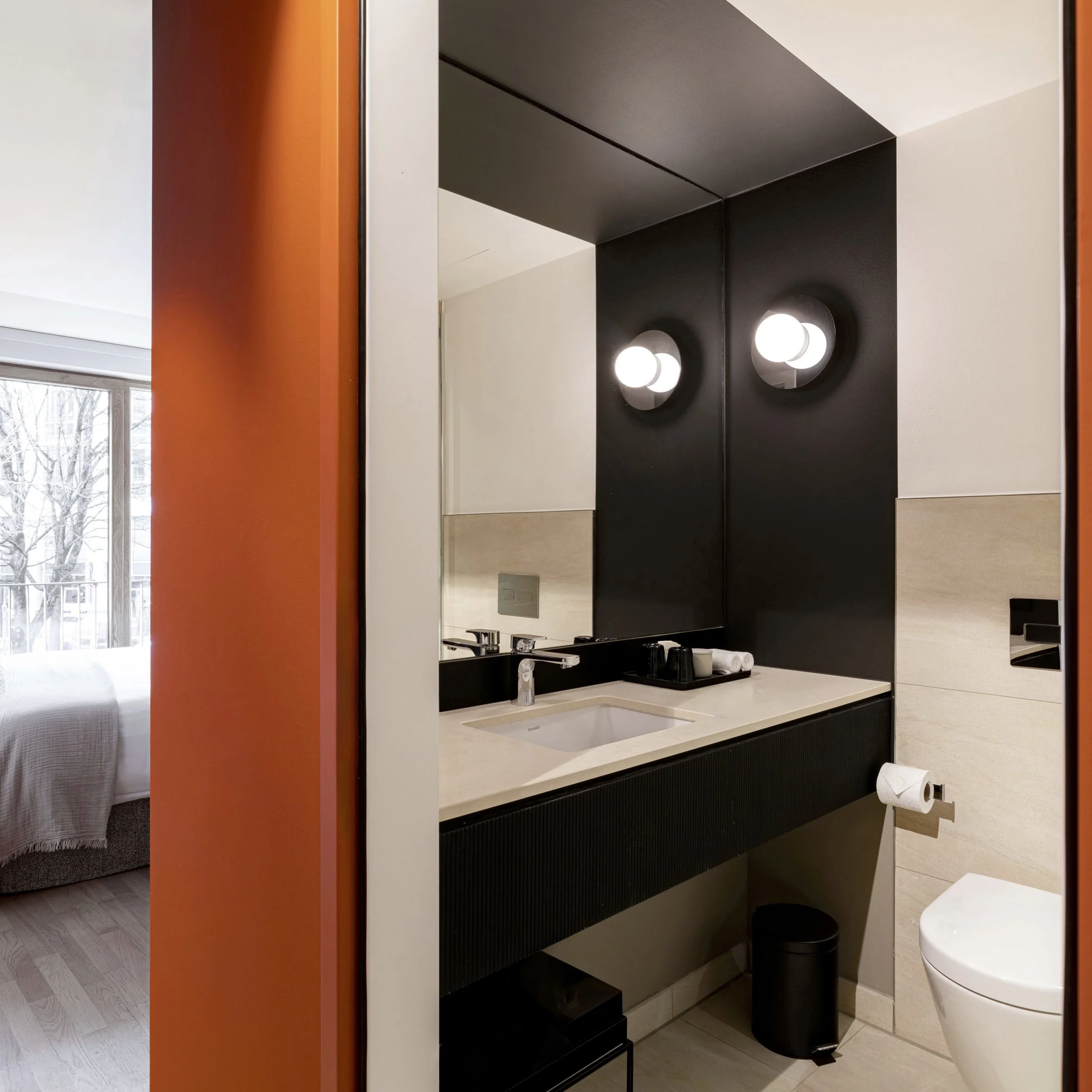 Moderne Badezimmer mit schwarzem Waschtisch, Spiegel und zwei runden Wandleuchten, weiße Dusche, orange Wand, Fenster mit Blick nach draußen