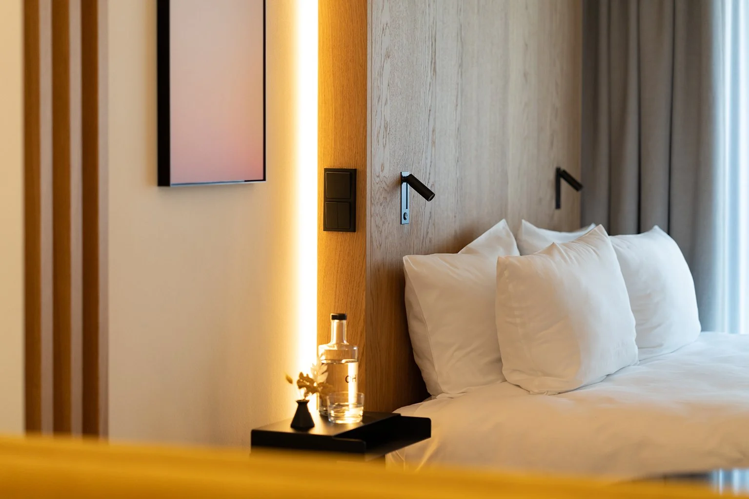 Schön angerichtetes Hotelzimmer mit weißem Bett, schwarzen Nachttisch, Wasserflasche, Gläser und Blumen, Holzwand und Vorhängen.