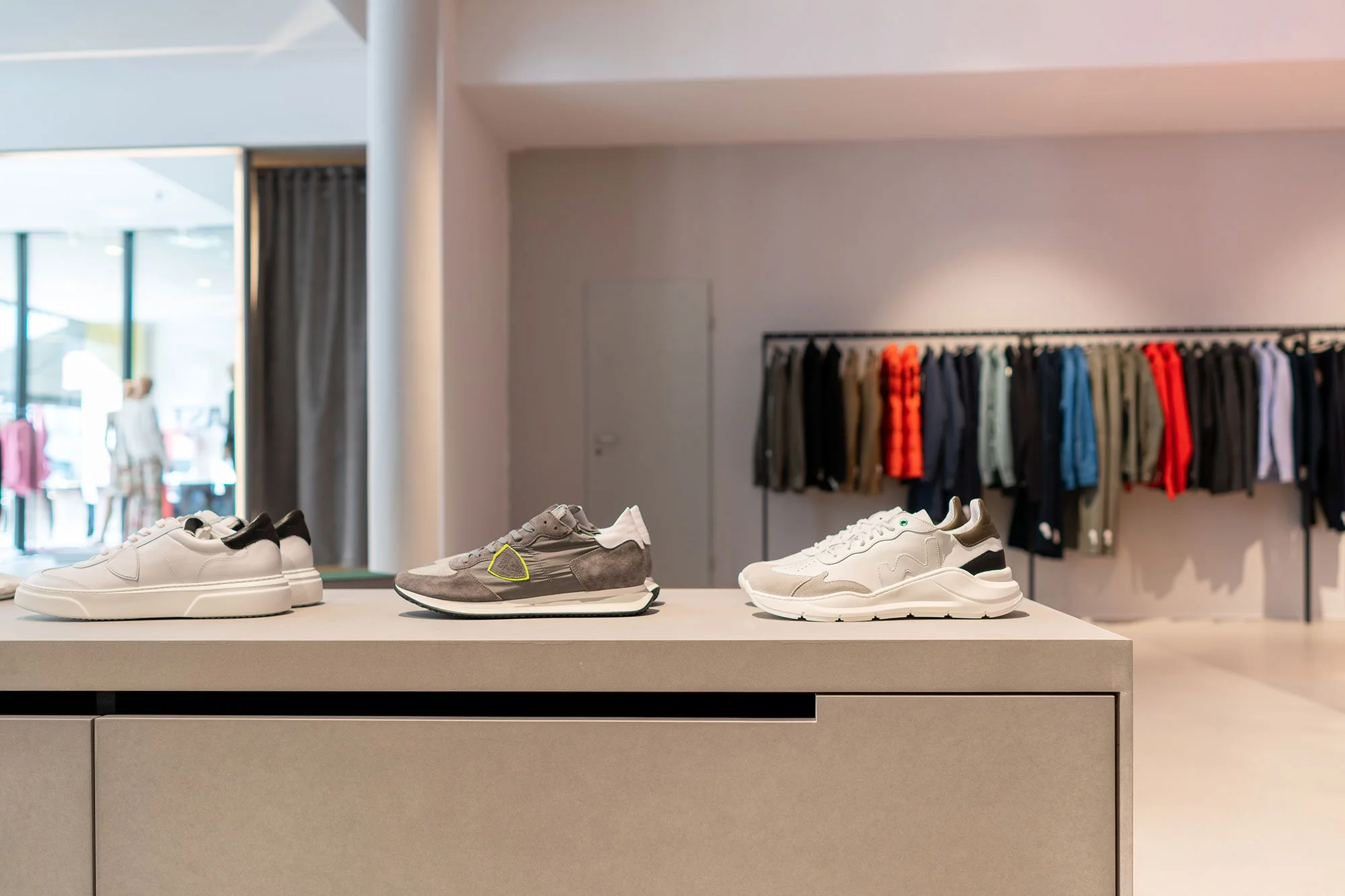 Drei Paar Turnschuhe auf einer weißen Verkaufstheke in einem Modegeschäft, im Hintergrund Kleidung auf einer Wand. Links eine große Glasscheibe mit Blick ins Einkaufszentrum.