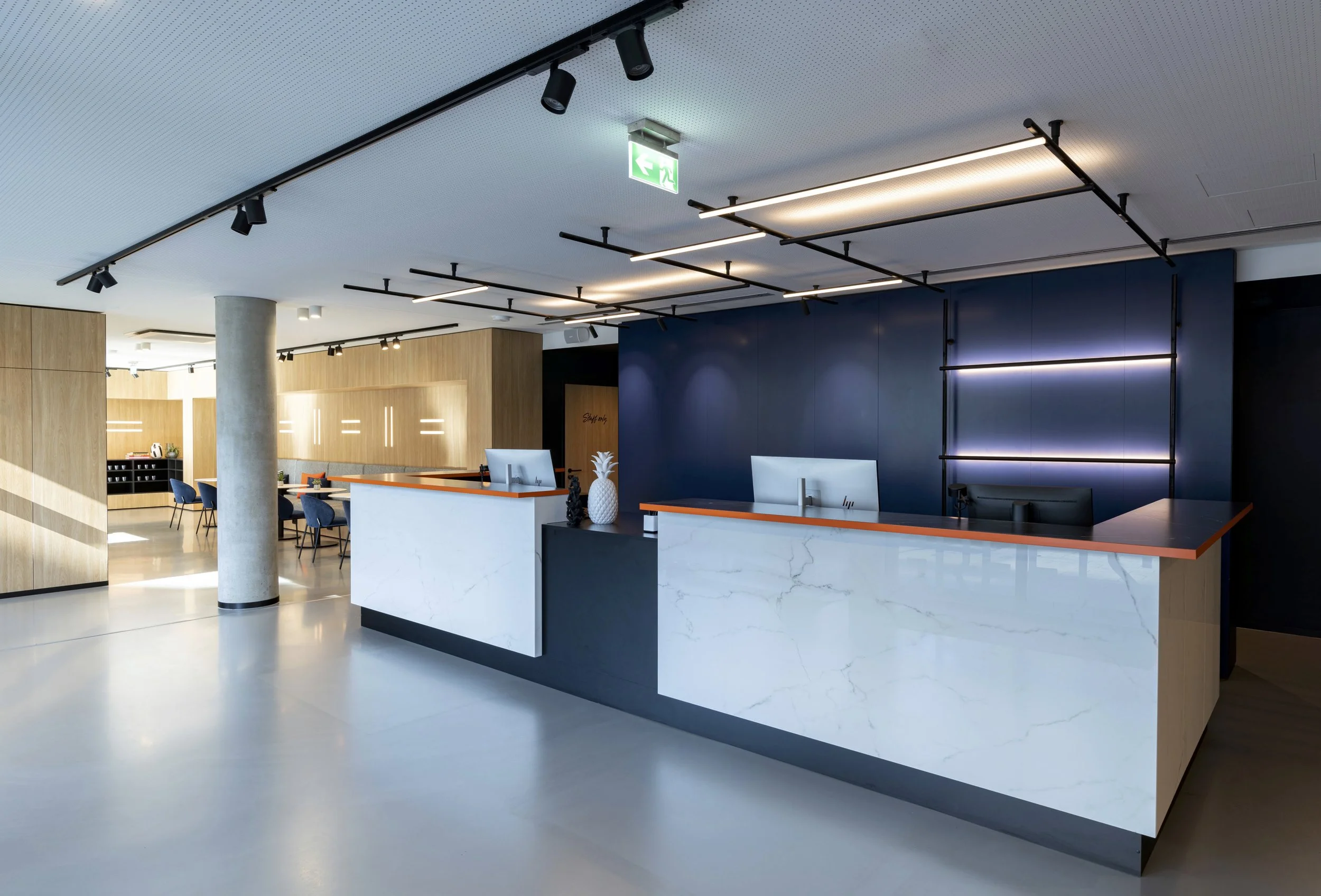 Moderne Lobby mit Empfangstresen aus weißem Marmor, blauer Wand und Sitzgelegenheiten. Deckenbeleuchtung und Notausgangsschilder hängen an der Decke.