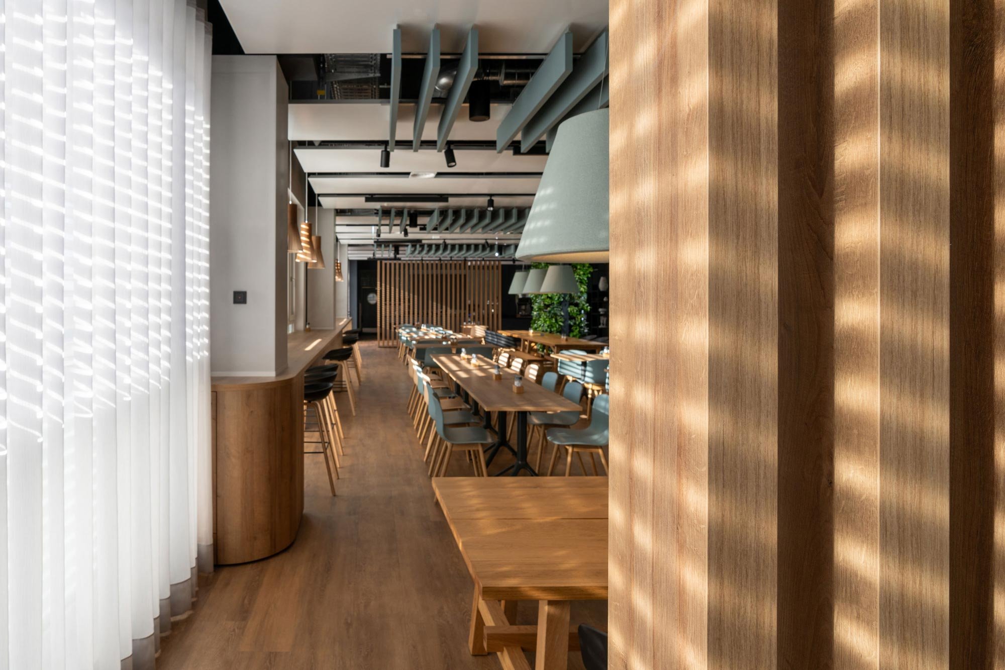 Innenraum eines modernen Restaurants mit Holztischen und Stühlen, deckenabhängige Beleuchtung und begrünte Wand im Hintergrund.