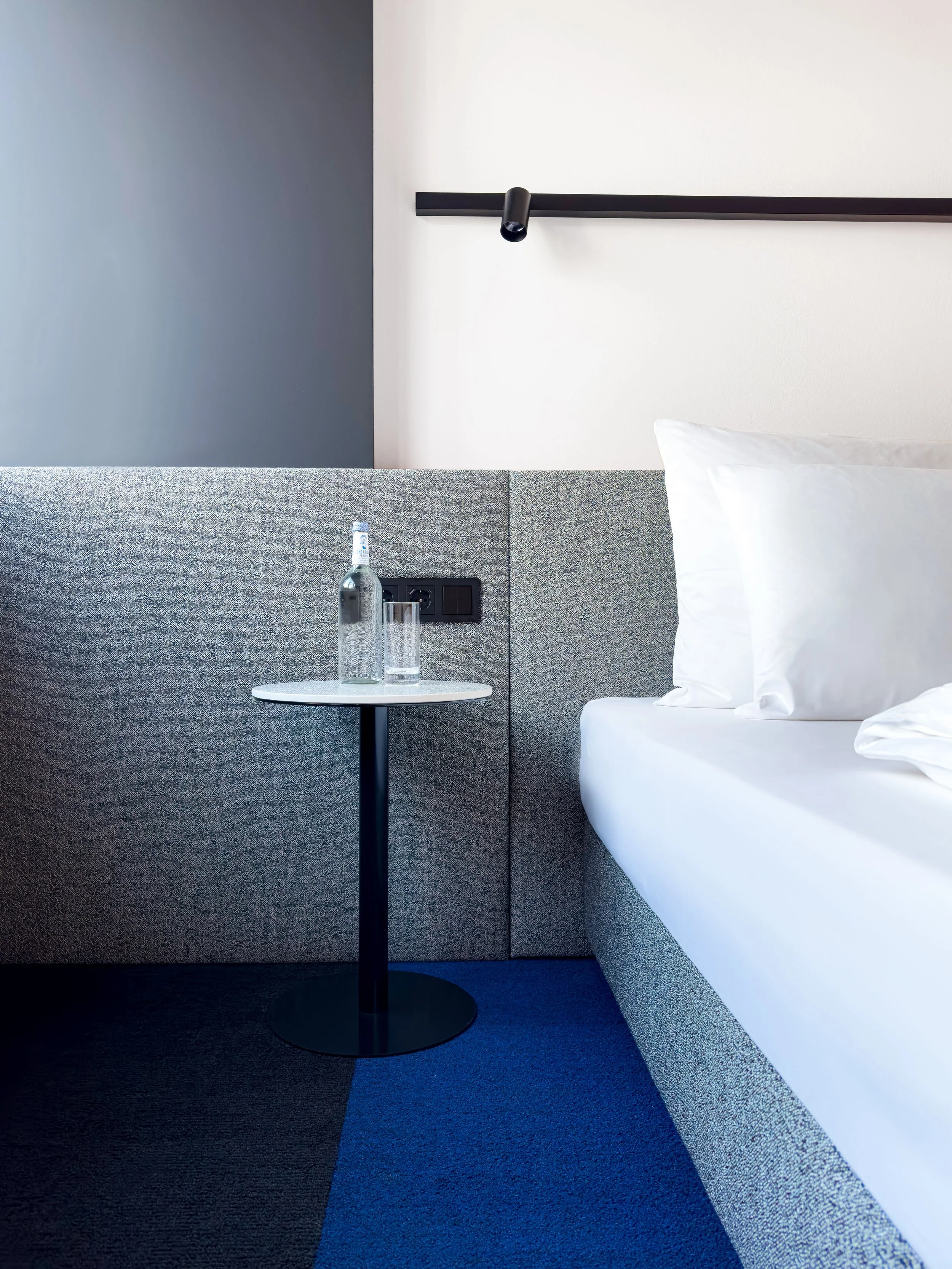 Ein moderner Hotelzimmerbereich mit grauem und weißen Wänden, einer Nachttischlampe, einem Wasserkocher auf einem kleinen Tisch, einem Glas Wasser und einer weißen Bettdecke.