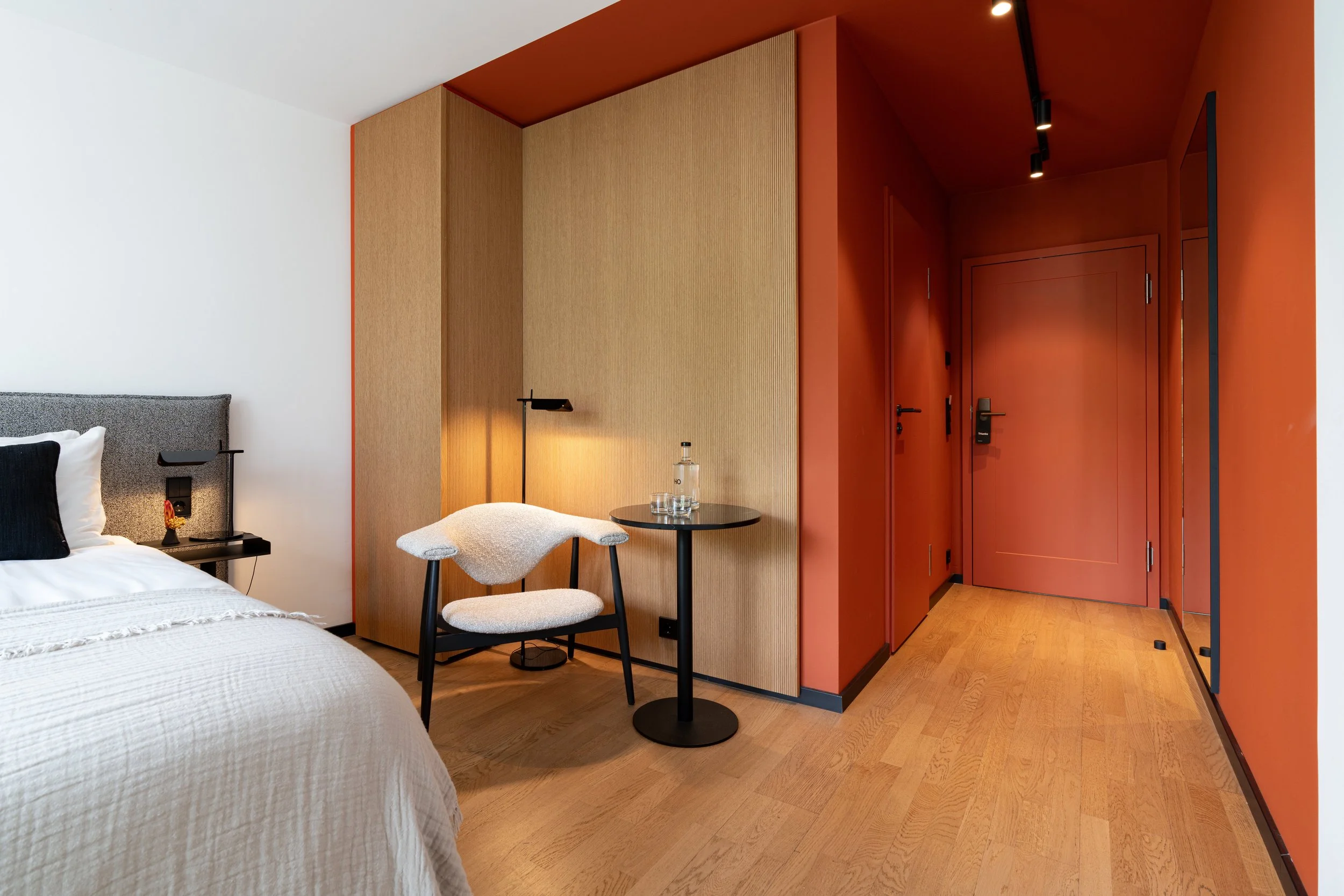 Modernes Hotelzimmer mit offenem Eingang, Holzfußboden, Bett mit grauem Kopfteil, schwarzem Nachttisch, Tischlampe, weißen Bettwäsche, einem Stuhl mit weißen Polster und runder schwarzen Tisch, rote Wände und Tür.