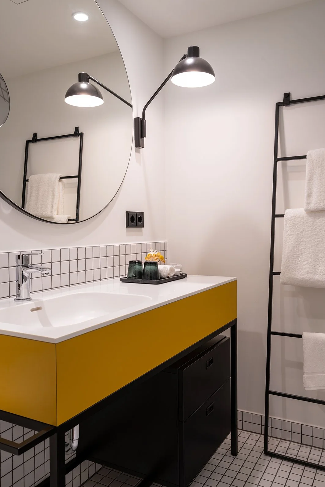 Moderner Badezimmerbereich mit weißem Waschtisch, gelber Schublade, schwarzem Unterschrank, großem runden Spiegel, Wandlampe, Handtuchheizkörper und kleinen quadratischen Fliesen.