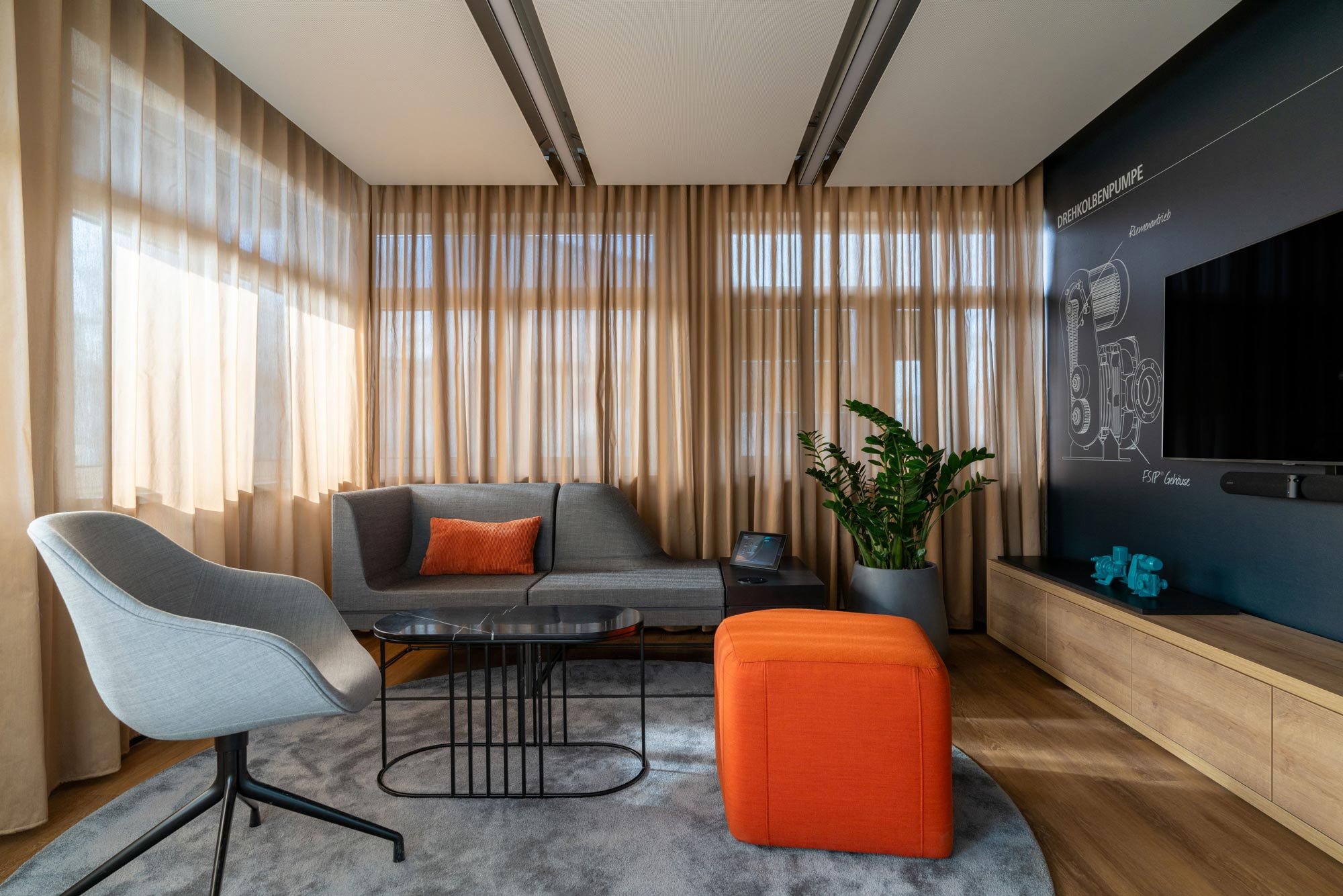 Ein modernes Wohnzimmer mit beigen Vorhängen, einem grauen Sofa, einem kleinen schwarzen Beistelltisch, einem weißen Armstuhl mit grauem Kissen, einem orangefarbenen Hocker, einer Pflanze in einem Topf, einem Fernseher an einer blauen Wand mit techni