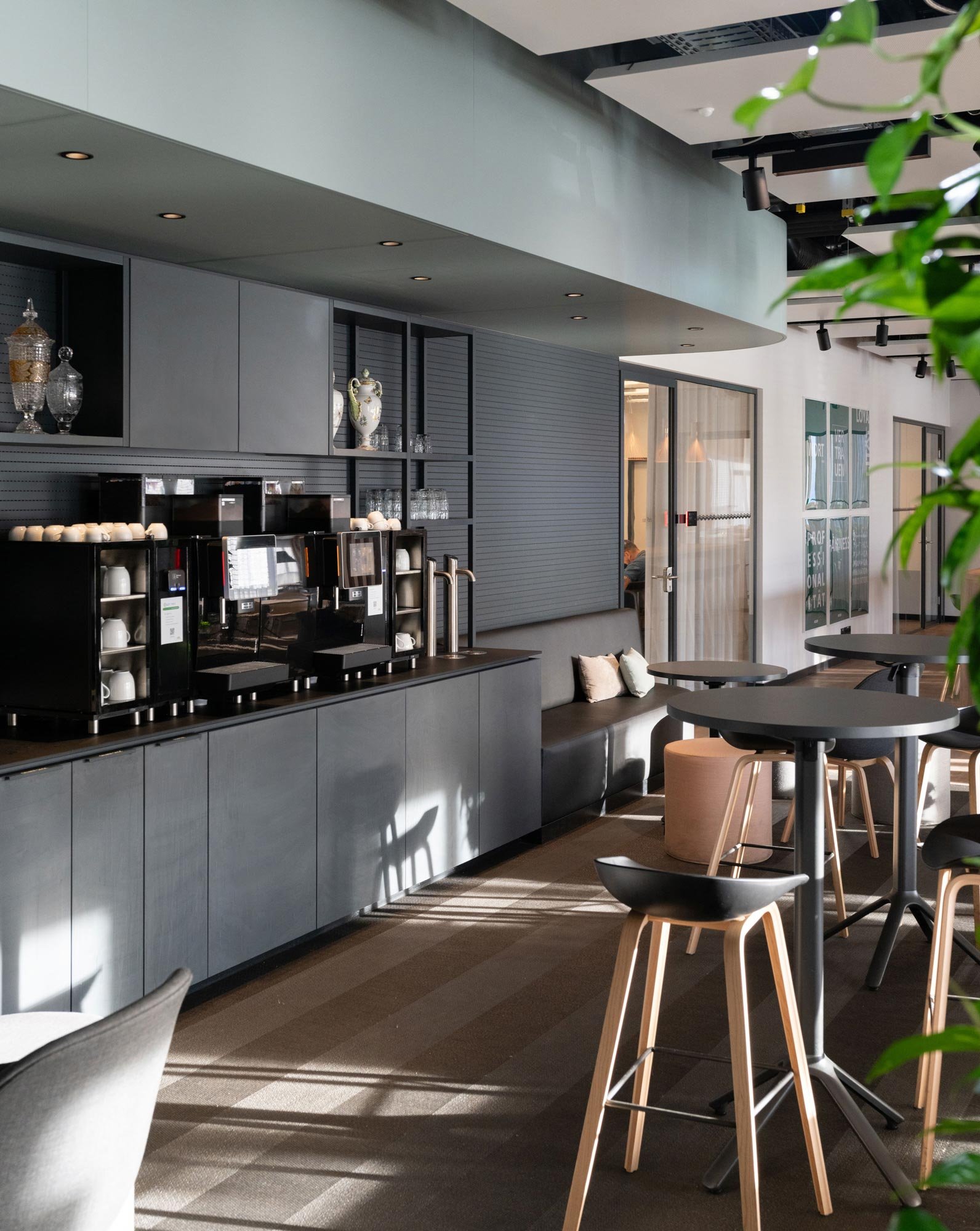 Innenaufnahme eines modernen Cafés mit schwarzem Kaffeeautomaten, schwarzen Tischen, Stühlen und einer schwarzen Wand mit Dekorvasen. Das Licht fällt durch große Fenster auf den Boden, es gibt Grünpflanzen im Vordergrund.