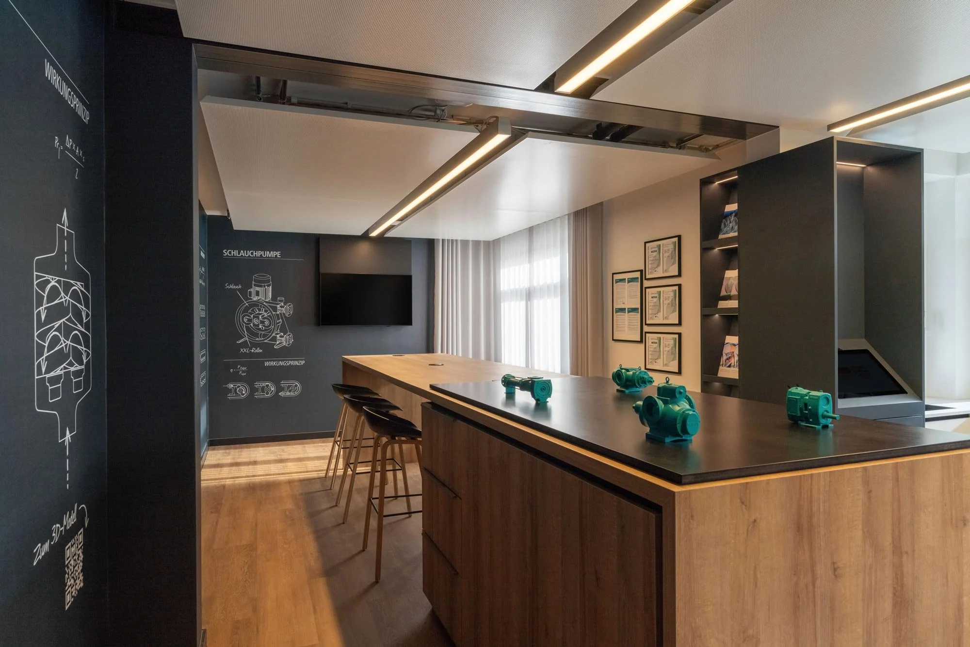 Innenraum eines modernen Schulungsraums mit einem großen Holztisch, mehreren Stühlen, bläulichen technischen Geräten auf dem Tisch, einer schwarzen Wand mit technischen Zeichnungen und Beschreibungen, einem Flachbildfernseher, großen Fenstern mit wei