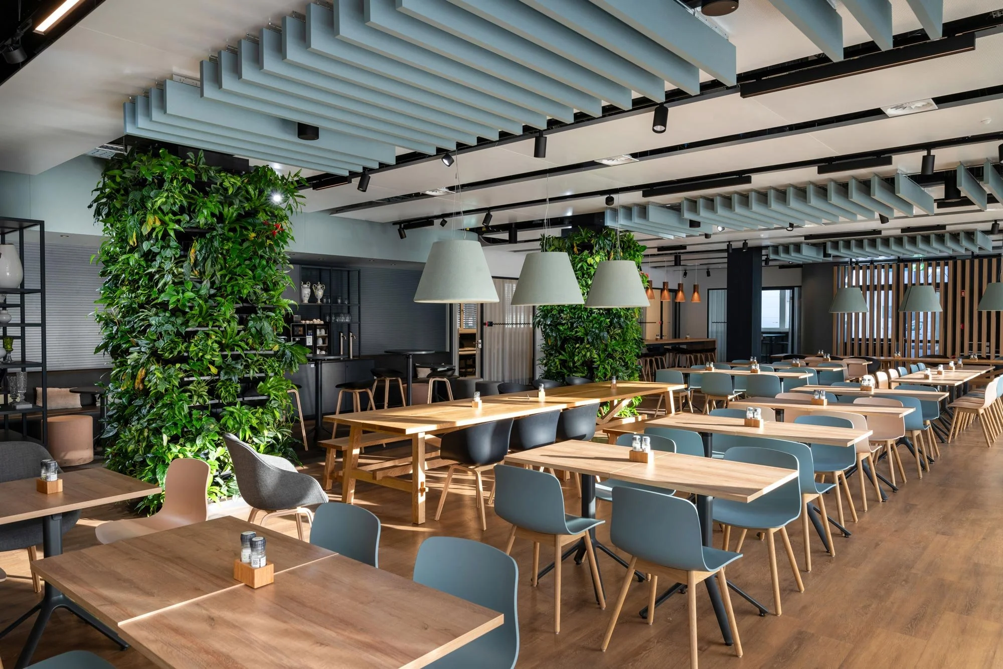 Ein modernes, ruhiges Restaurant mit mehreren leeren Tischen und Stühlen, viel Grünpflanzen, schützende Deckenleisten an der Decke und große Lampen für angenehmes Licht.