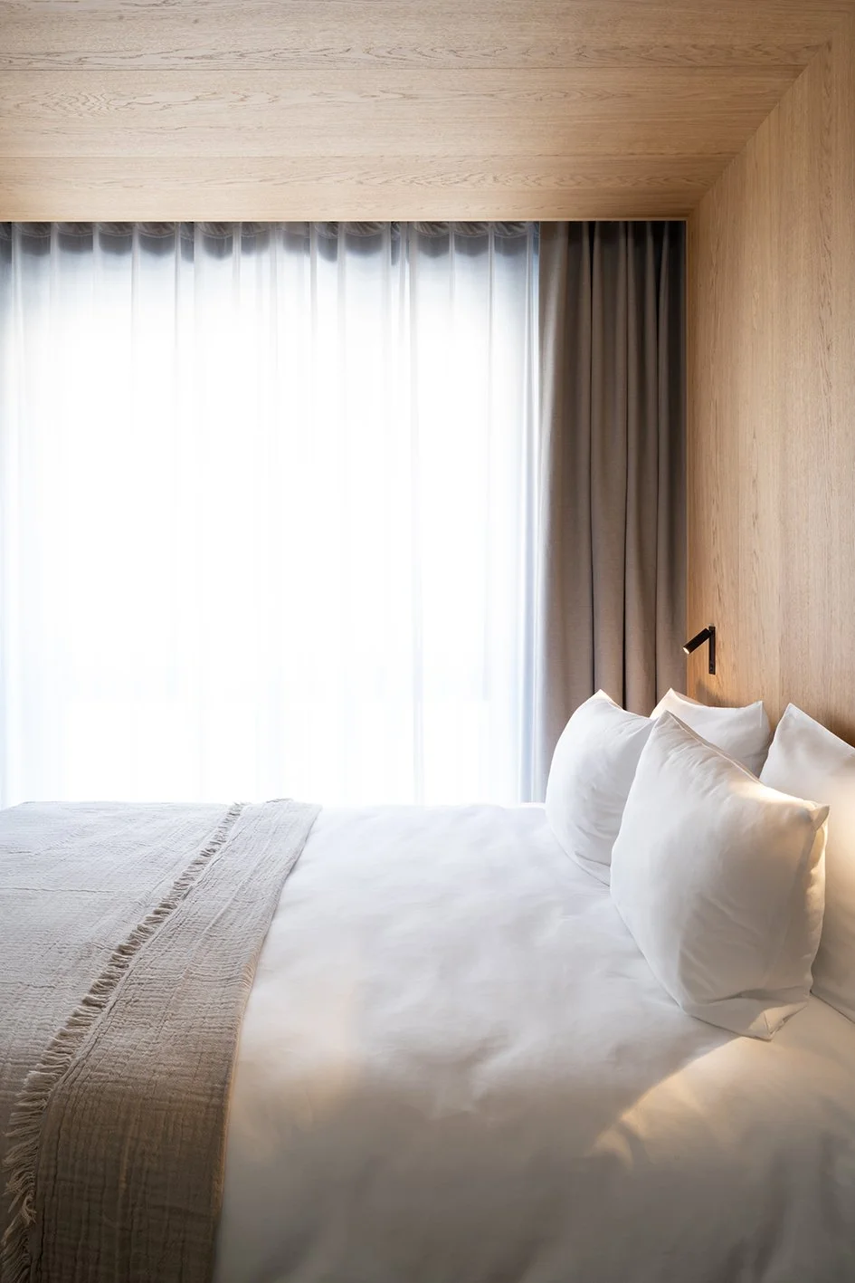 Ein modernes Schlafzimmer mit weißen Bettwäsche, mehreren Kissen, Holzwand und großen Fenstern mit weißen und grauen Vorhängen.