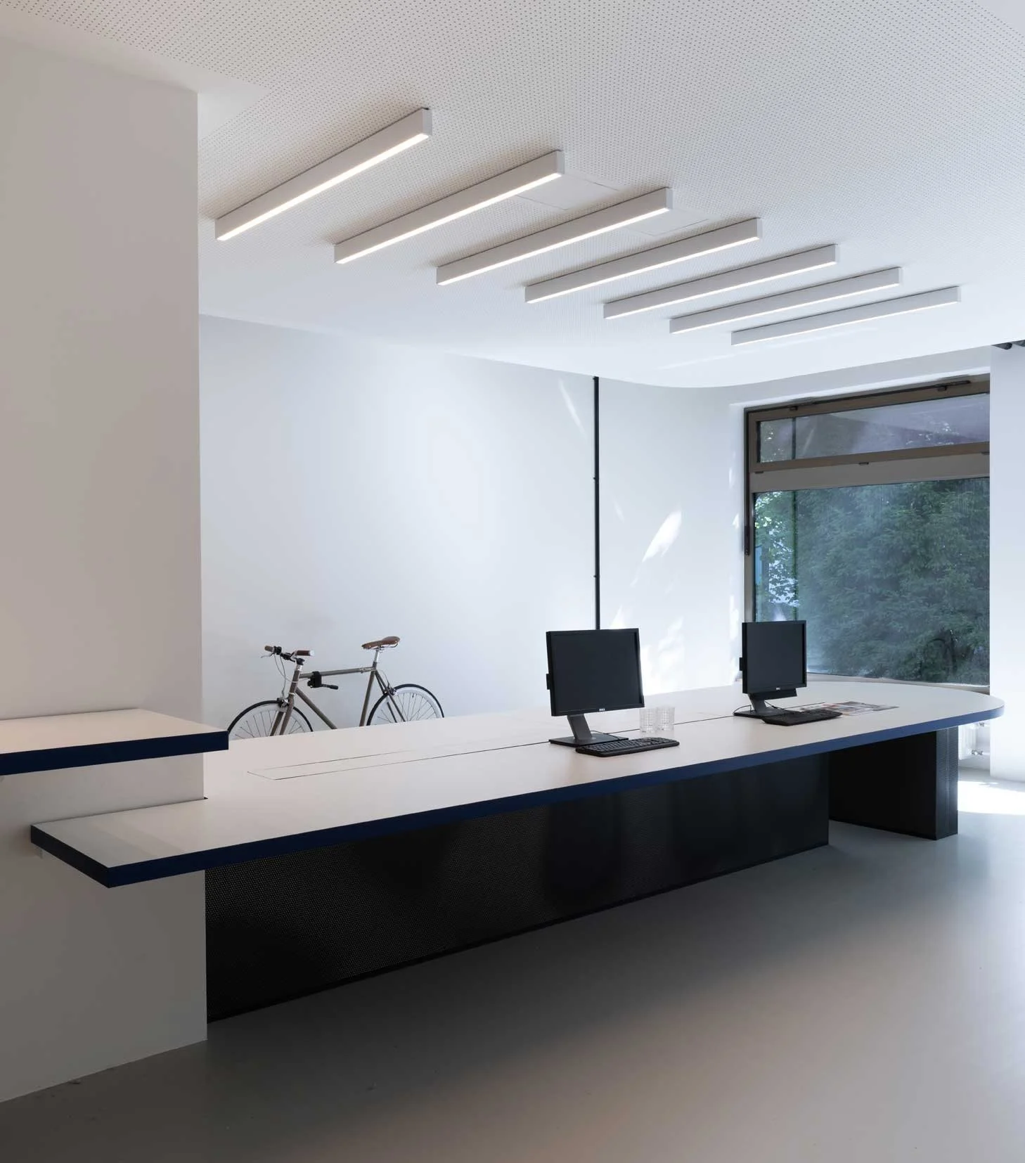 Ein moderner, heller Empfangsbereich mit weißen Wänden, einer langen Theke und zwei Computern. Im Hintergrund befindet sich ein großes Fenster mit Blick nach draußen und ein Fahrrad steht an der Wand.