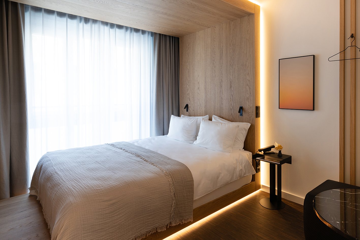 Modernes Hotelzimmer mit Bett, Nachttisch, Wandlampen, Kunstwerk, Vorhängen im Hintergrund und Holzwandgestaltung