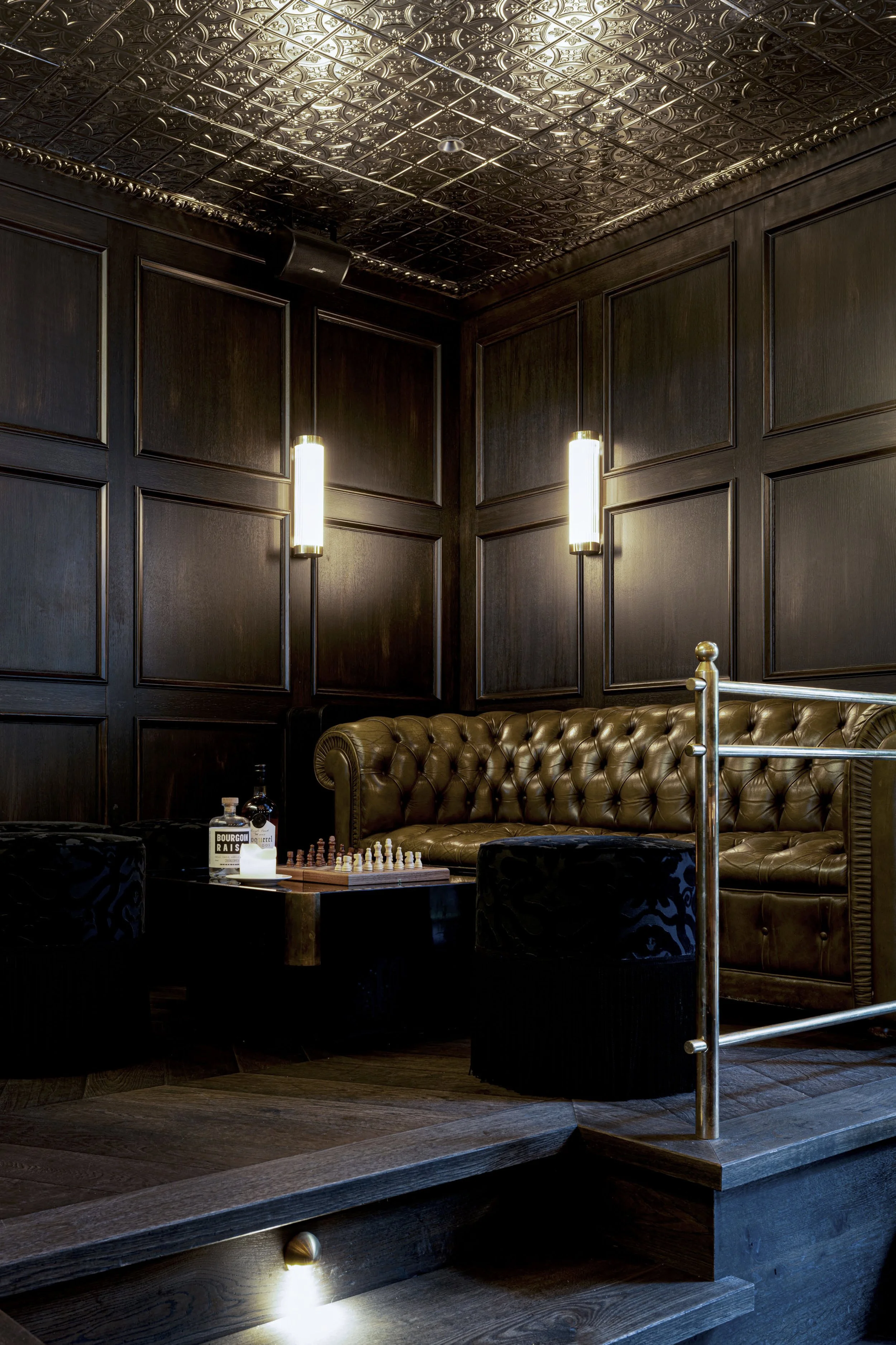 Ein gemütliches Lounge mit dunklen Holzpaneelen an den Wänden, einer braunen Ledercouch, einem Tisch mit Schachspiel und Alkoholflaschen, beleuchtet von Wandlampen mit modernem Design, auf einem dunklen Holzboden.