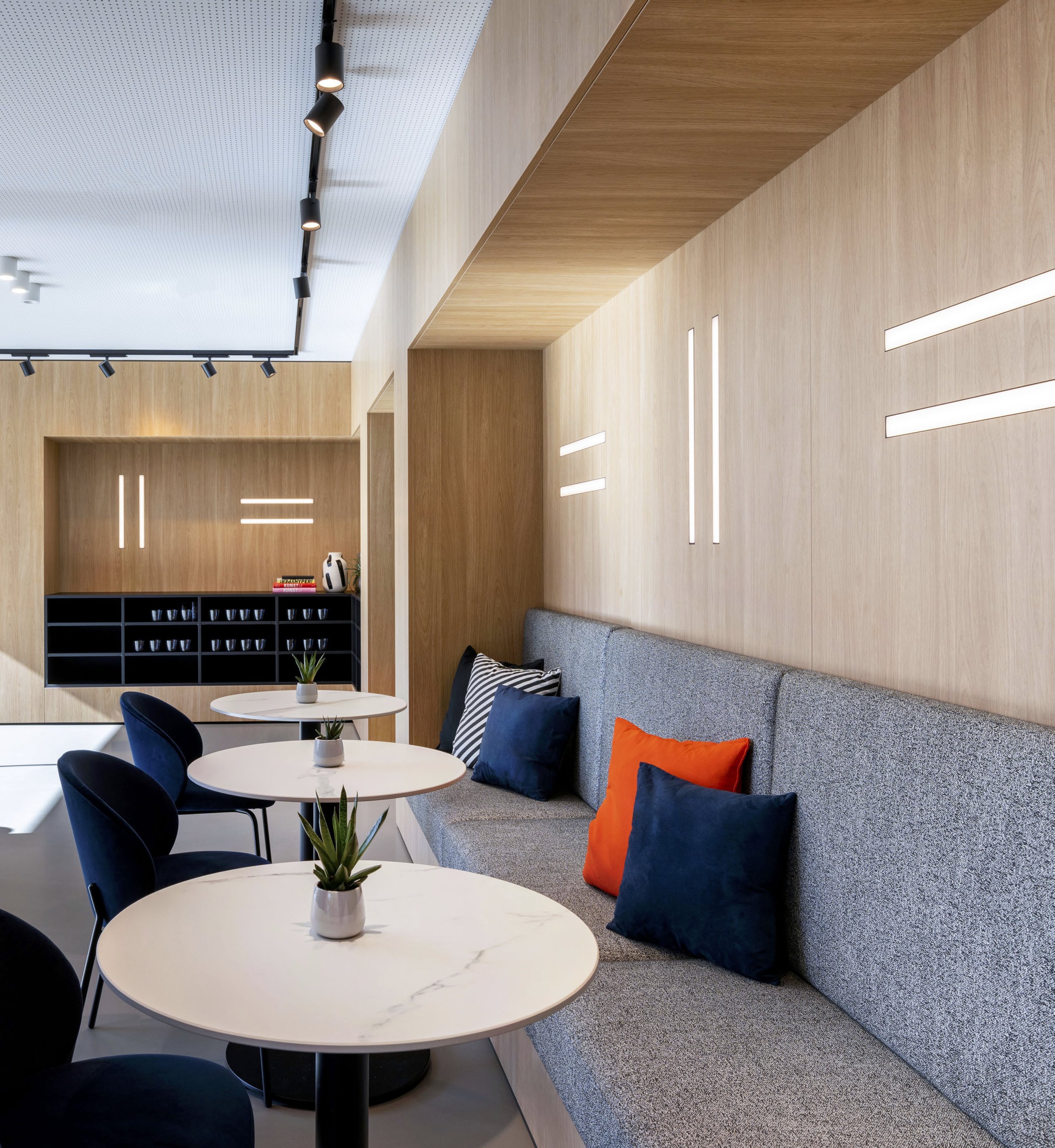 Moderne Lounge mit weißen Tischen, blauen Stühlen, einer grauen Couch mit bunten Kissen, Holz-paneelierten Wänden und dekorativen Lichtleisten.