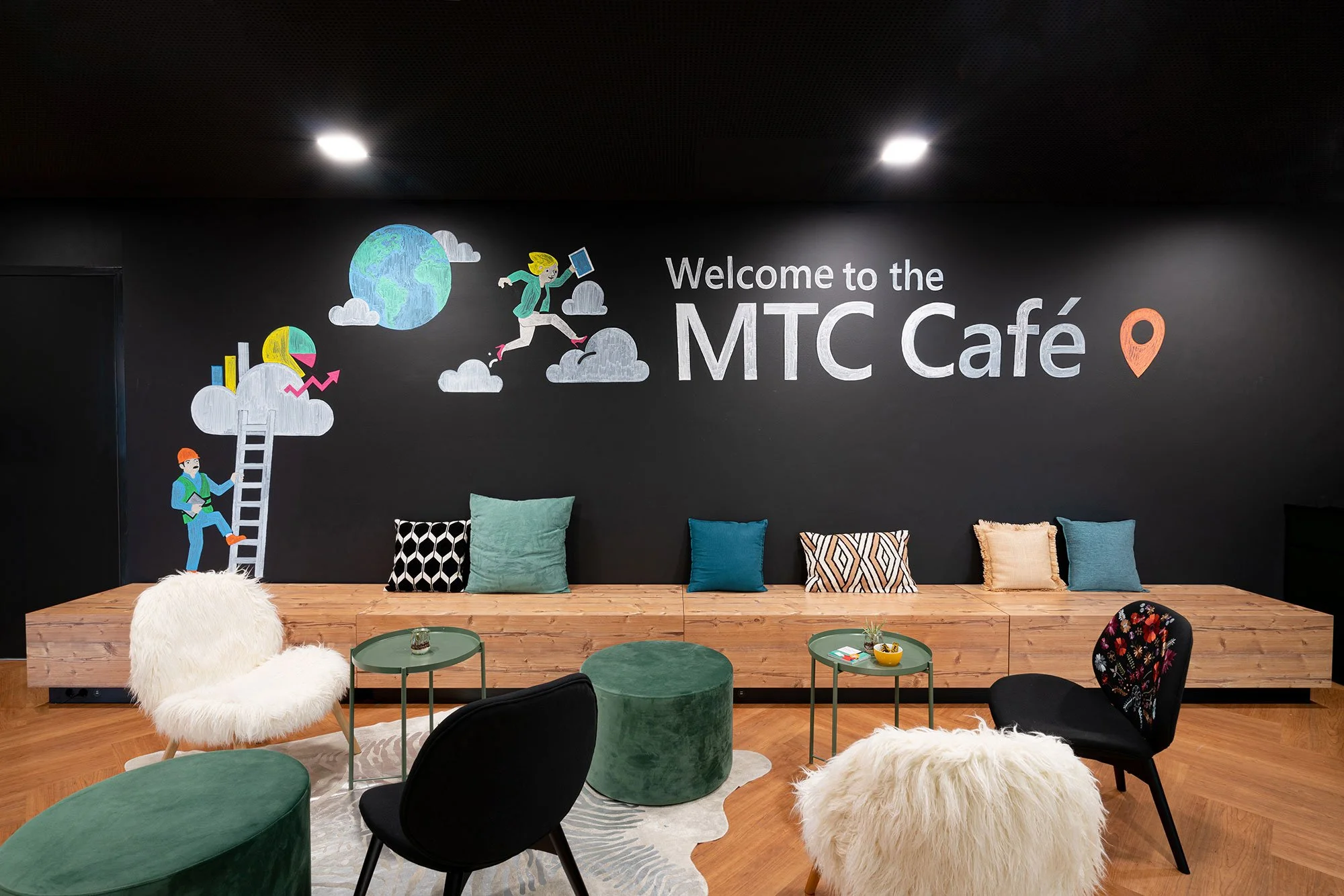 Bunte Sitzgruppen mit Kissen und einen Wanddekoration mit der Aufschrift Willkommen im MTC Café auf dunkler Wand. Darunter befinden sich gemusterte Kissen, ein nachhaltiger Holzbank und bequeme Sessel.