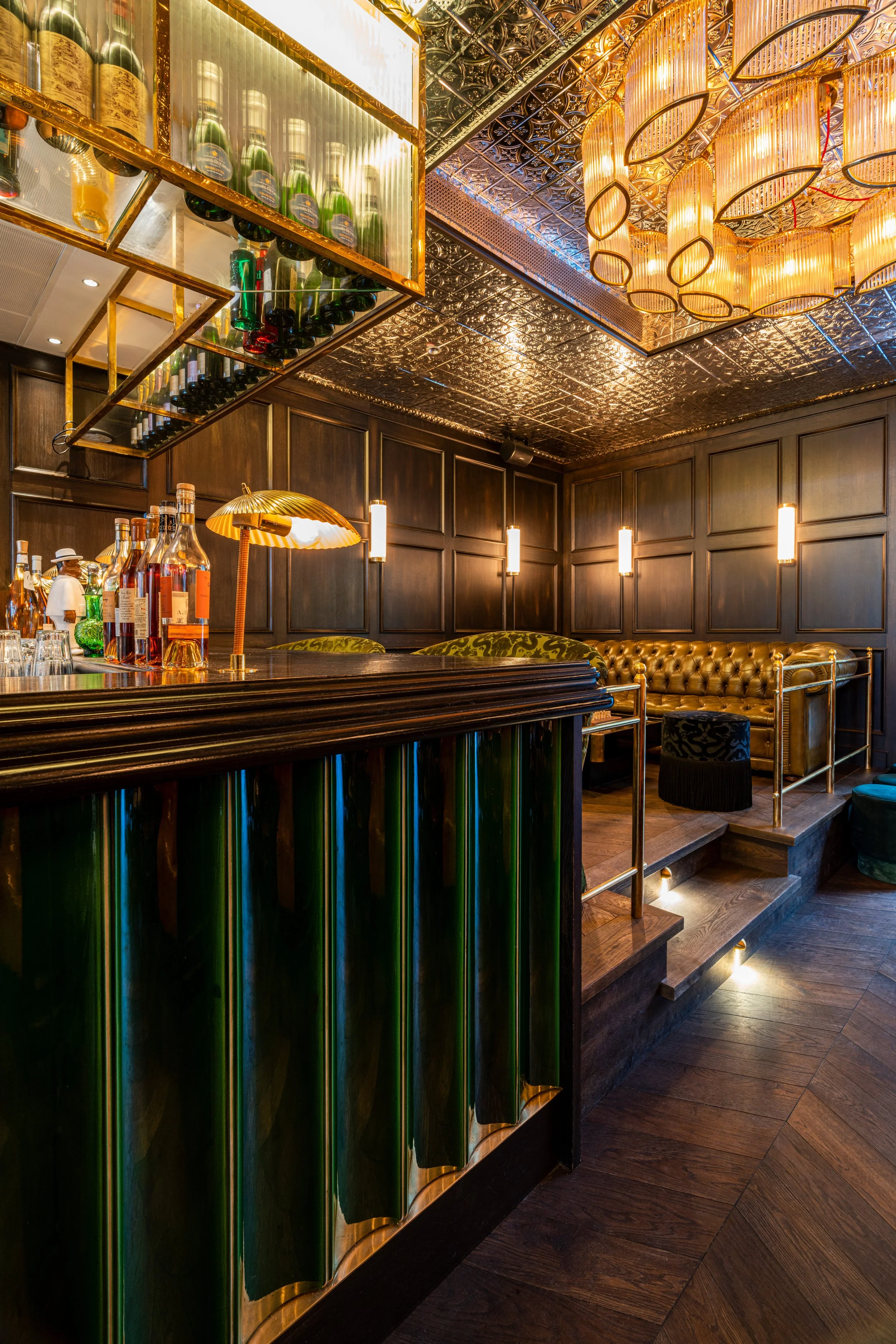 Elegante Bar mit dunklem Holz, goldfarbenen Akzenten, einer ansprechenden Beleuchtung und einer Bar mit verschiedenen Getränkeflaschen, darunter Wein, Spirituosen und Champagner; im Hintergrund stilvolle Sitzgelegenheiten und eine dekorative Decke.