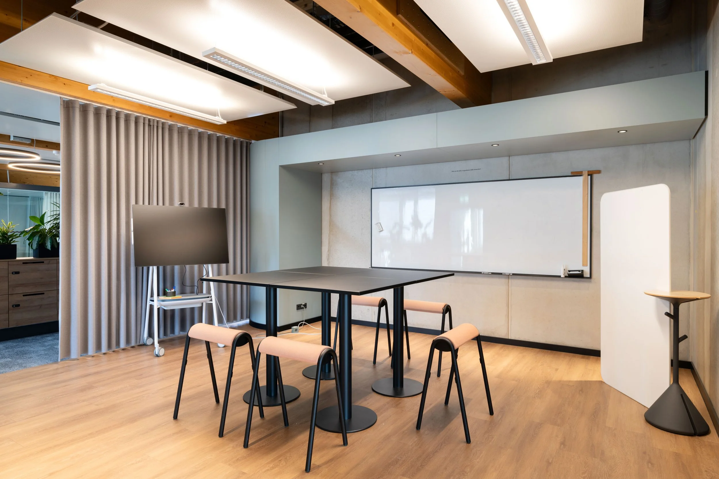 Moderner Konferenzraum mit Whiteboard, digitalen Monitor, Tischen und Stühlen, mit Holzbalken und Vorhängen.