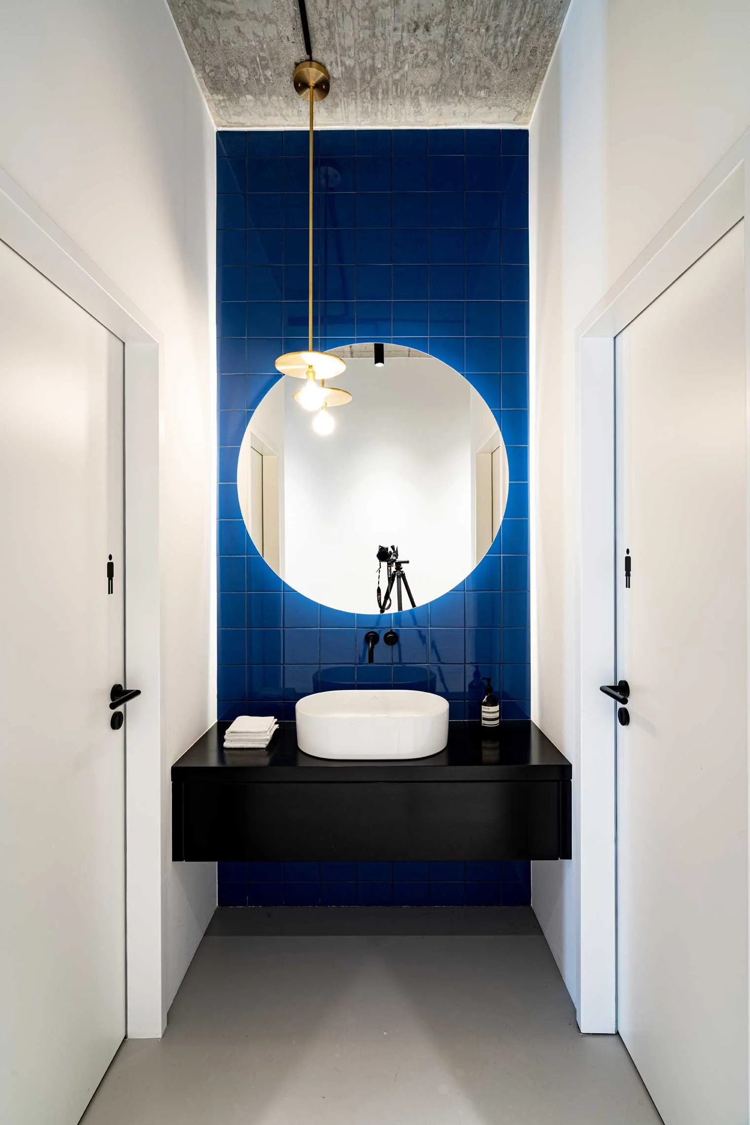 Ein modernes, minimalistisch eingerichtetes Badezimmer mit blauer Fliesewand im Spiegel, eine schwarze Waschtischplatte mit ovalem Waschbecken, Spiegel, Deckenleuchte, Handtüchern und einem Fotoapparat auf einem Stativ im Spiegel