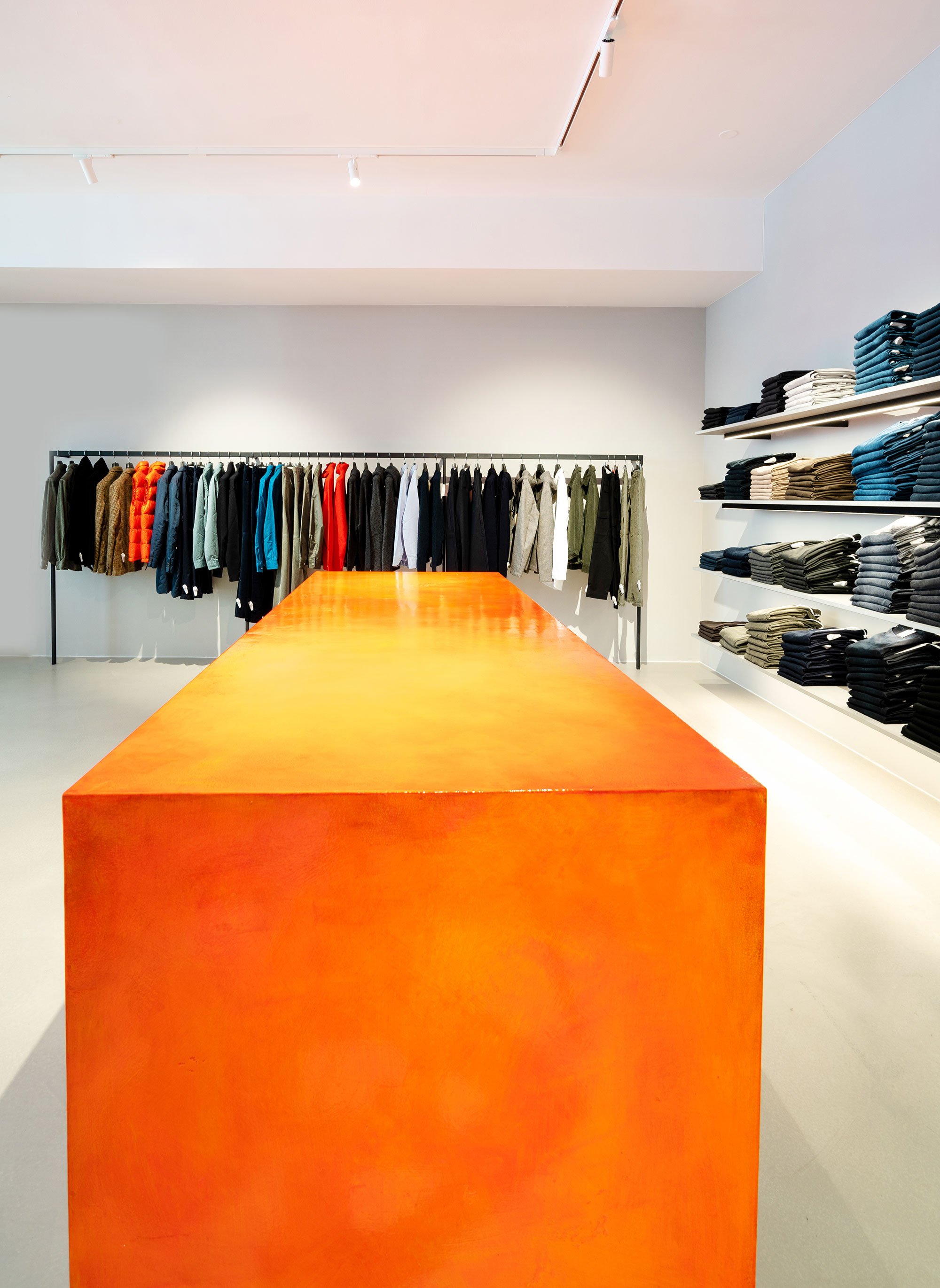 Ein Kleidungsgeschäft mit einer Kleiderstange voller Jacken und Pullovern sowie Regalen mit gefaltenen Hemden und Hosen. Im Vordergrund ein orangefarbener Tisch.