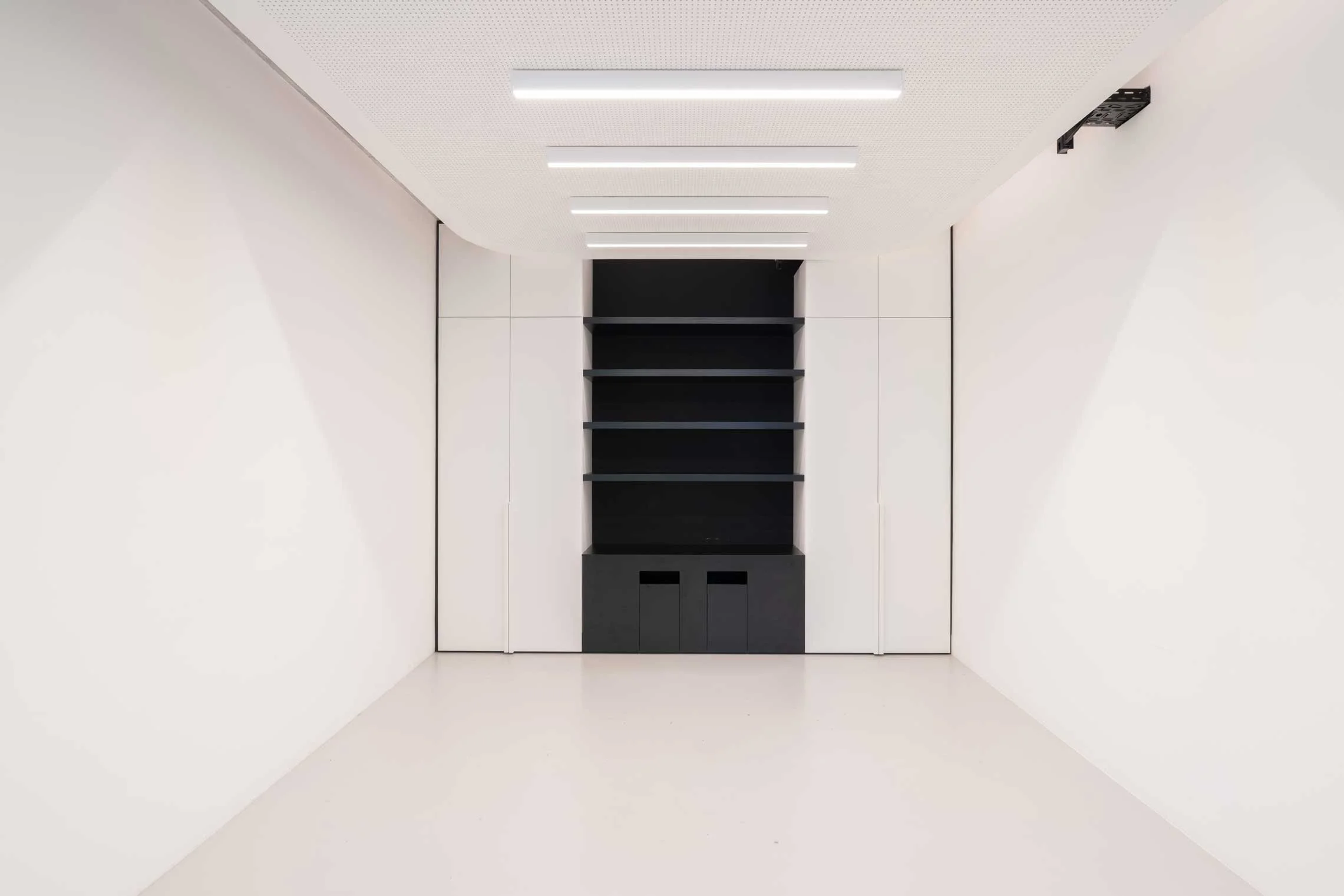 Ein modern gestalteter Raum mit weißen Wänden, perforiertem weißen Deckenpaneel und integrierten Lichtleisten. Am Ende des Raums befindet sich eine schwarze Wand mit offenen schwarzen Regalen und geschlossenen weißen Schränken.