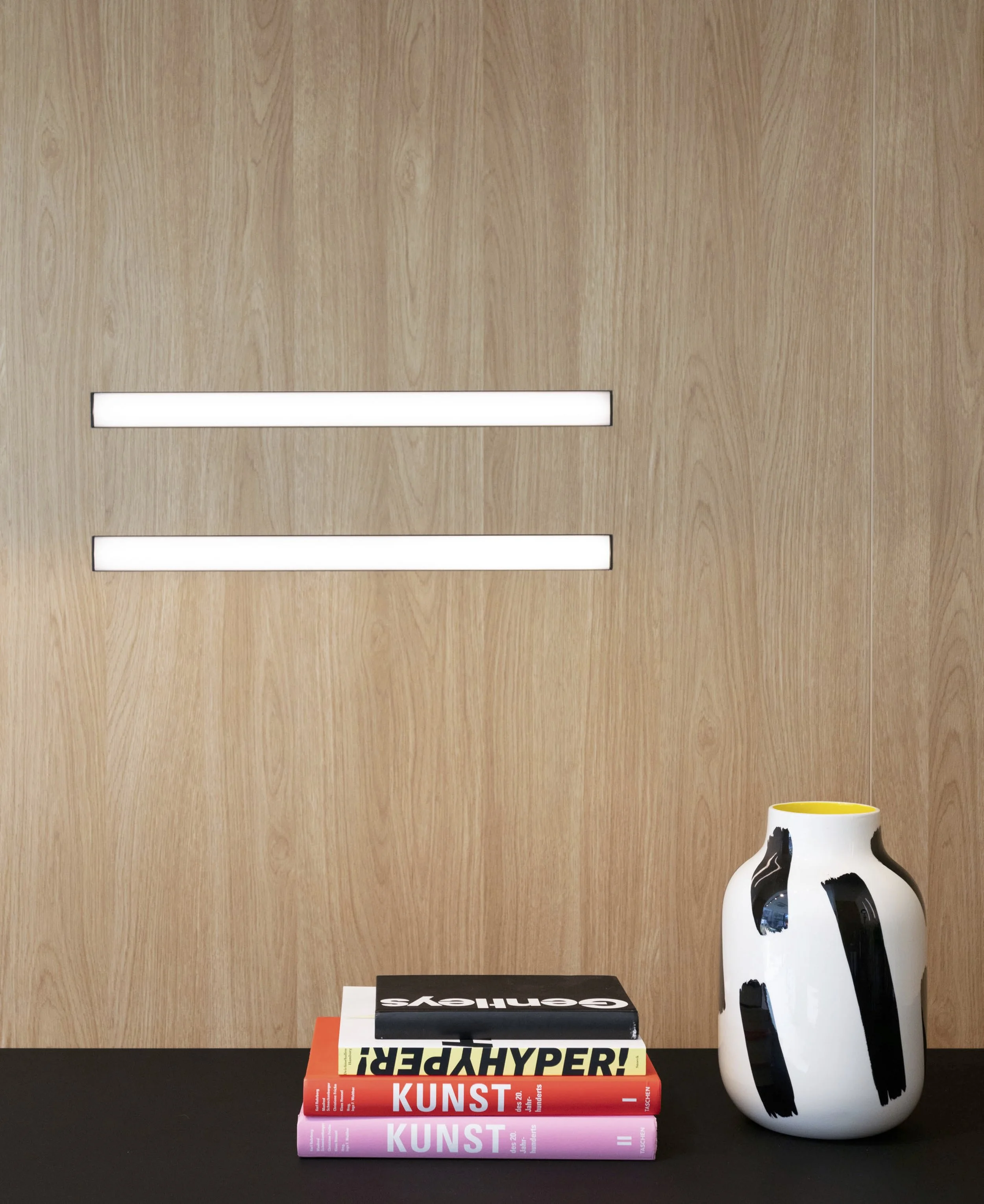 Ein schwarzer Tisch mit gestapelten Kunstbüchern, daneben eine weiße Vase mit schwarzen Malereien, im Hintergrund eine Holzvertäfelung.