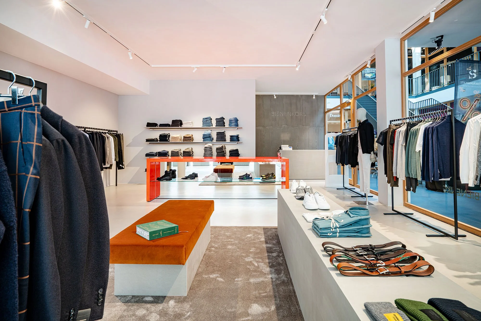 Innenansicht eines Modegeschäfts mit Kleidung, Schuhen und Accessoires, helle Beleuchtung, moderne Einrichtung, große Fenster, Produkte auf Tischen und Kleiderstangen.