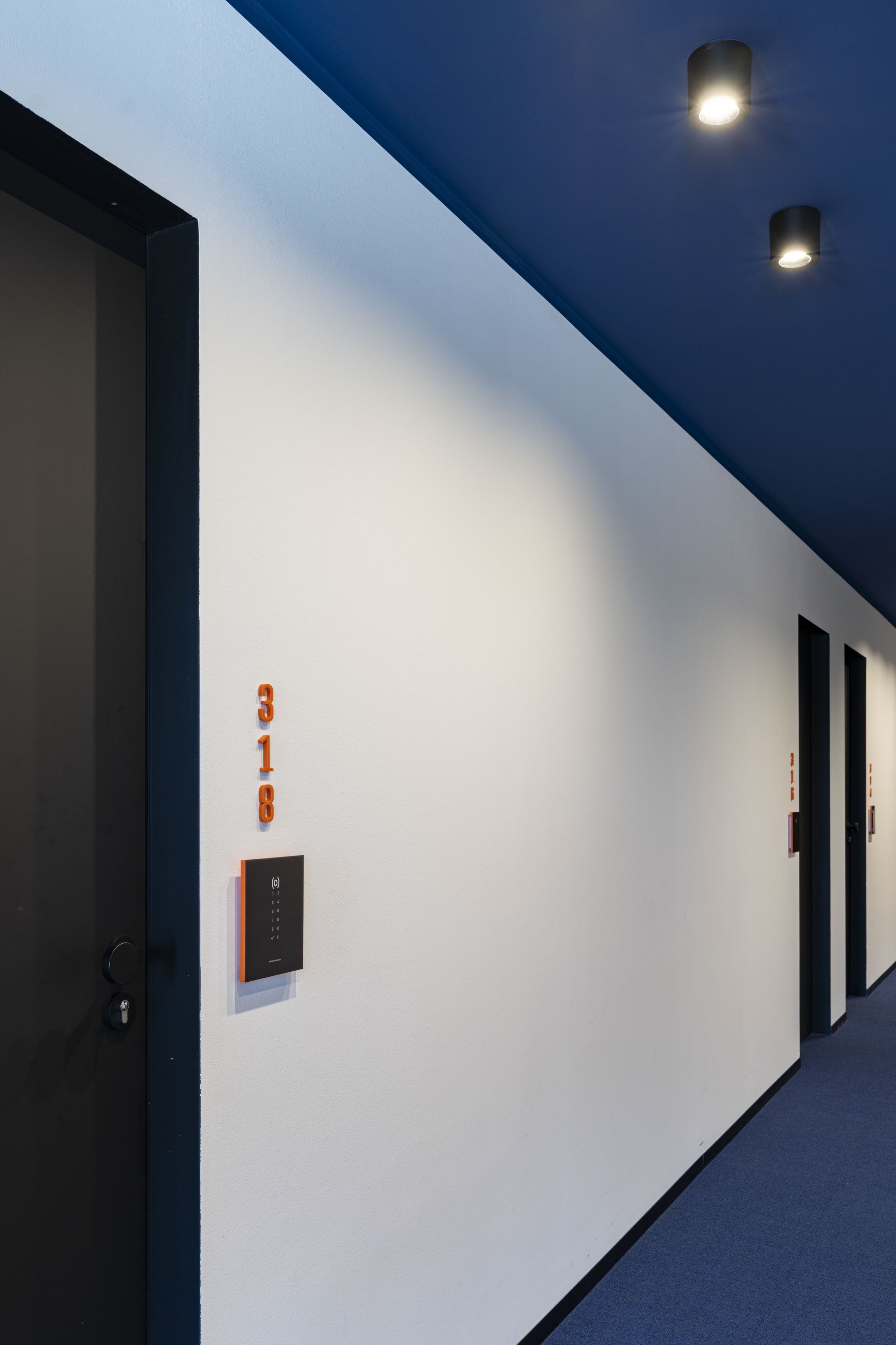 Innenaufnahme eines modernen Aufzugs mit dunkelblauer Decke, weißen Wänden und orangefarbenen Markierungen. Es gibt zwei schwarze, runde Deckenleuchten an der blauen Decke. Das Bild zeigt die linke Seite des Aufzugs mit einem schwarzen Türrahmen und 