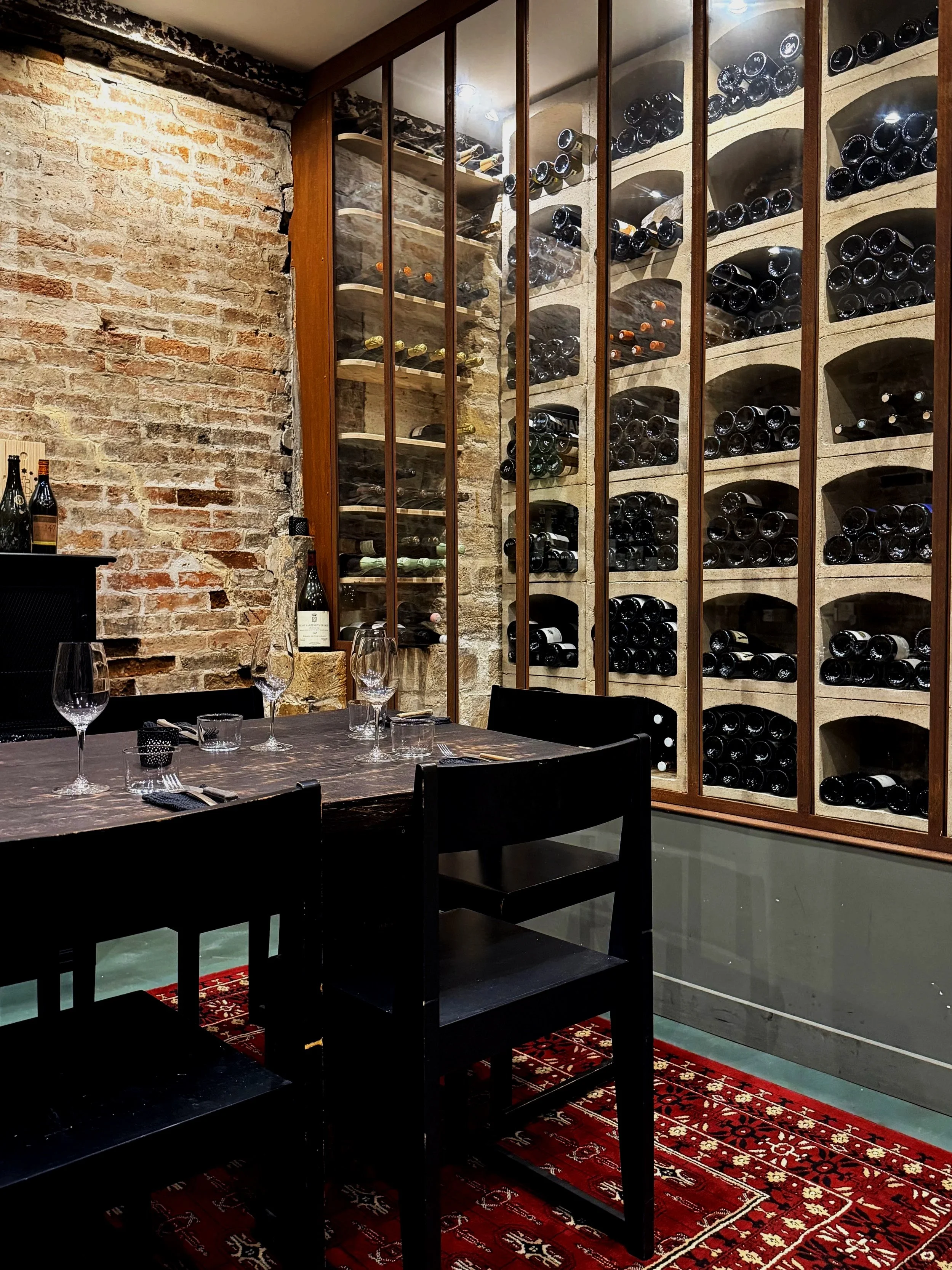 A rustic wine cellar with a brick wall on the left and a large wooden wine rack behind glass doors on the right, filled with numerous bottles of wine. In front of the rack is a dark wooden dining table set with wine glasses, some utensils, and a decorative rug underneath.