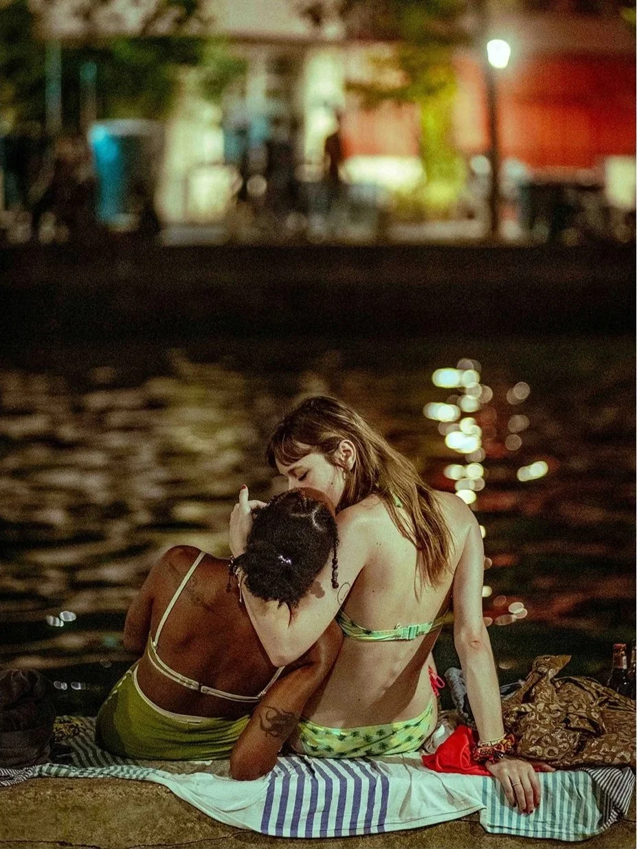Two women sitting near a body of water at night, embracing each other affectionately.