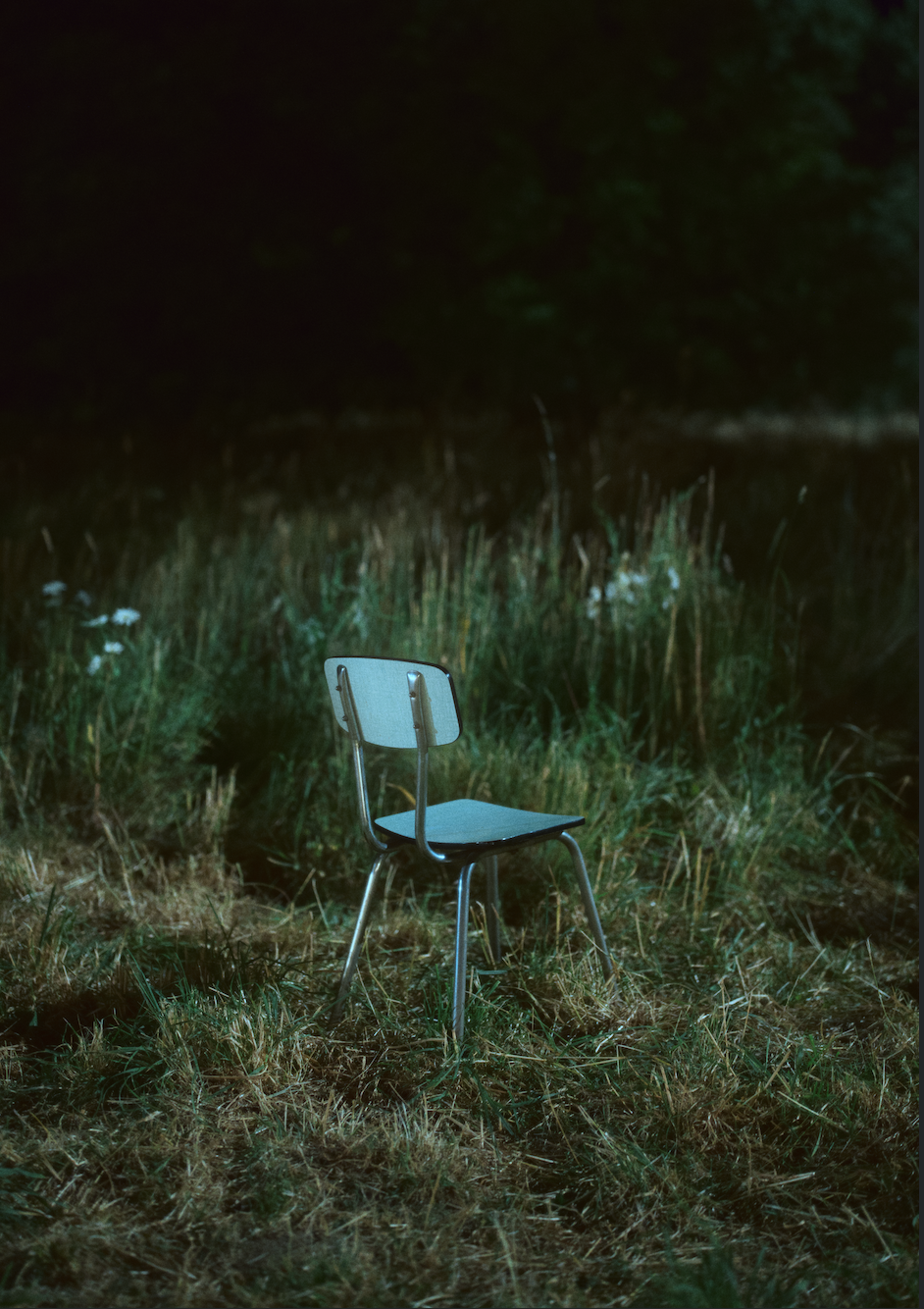 A blue metal and wood chair sits alone in a grassy field with trees in the background at dusk or night.